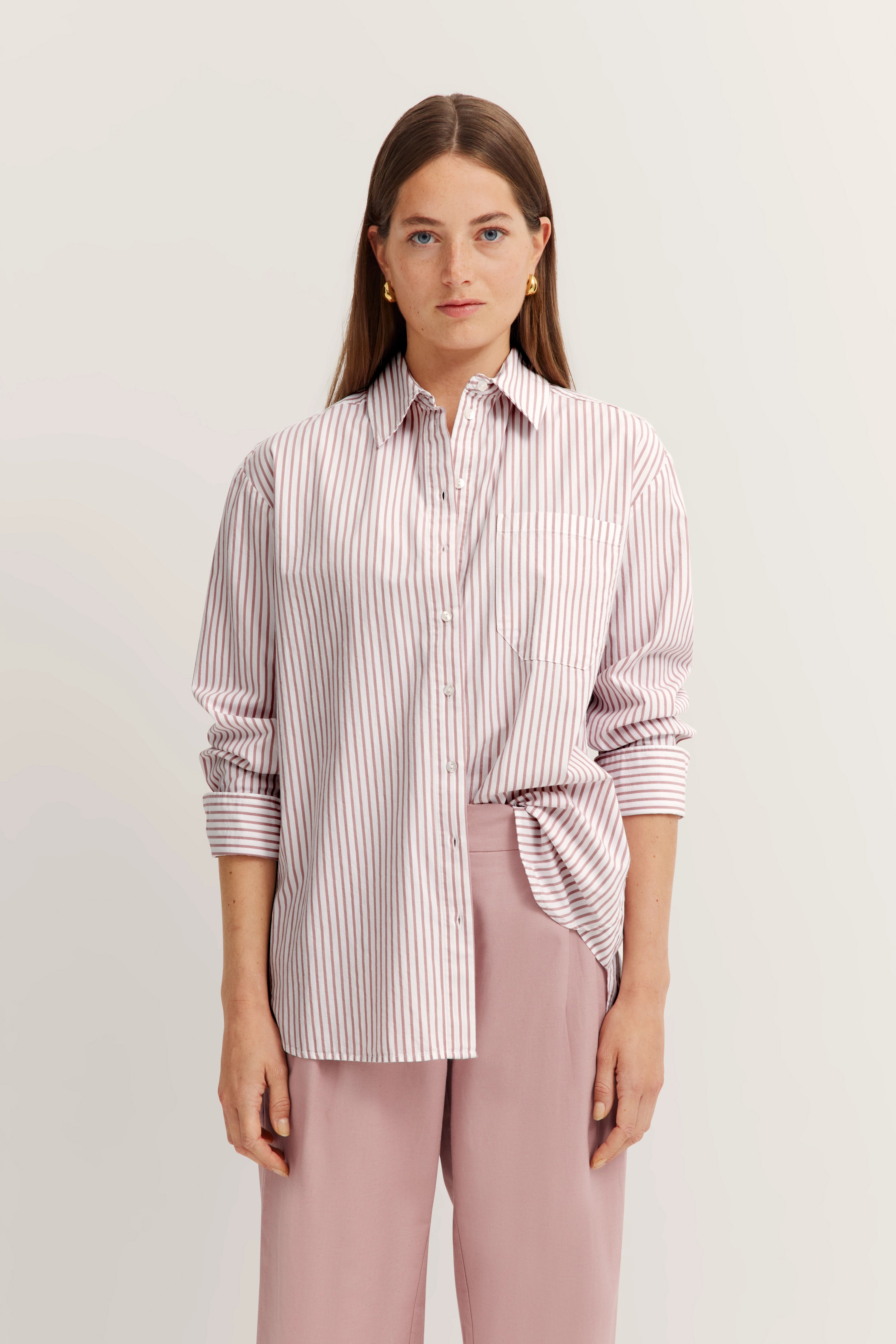 Gestreifte Bluse in Rosa-Weiß, locker über rosa Hose getragen, Modelbild, entspanntes Posing.