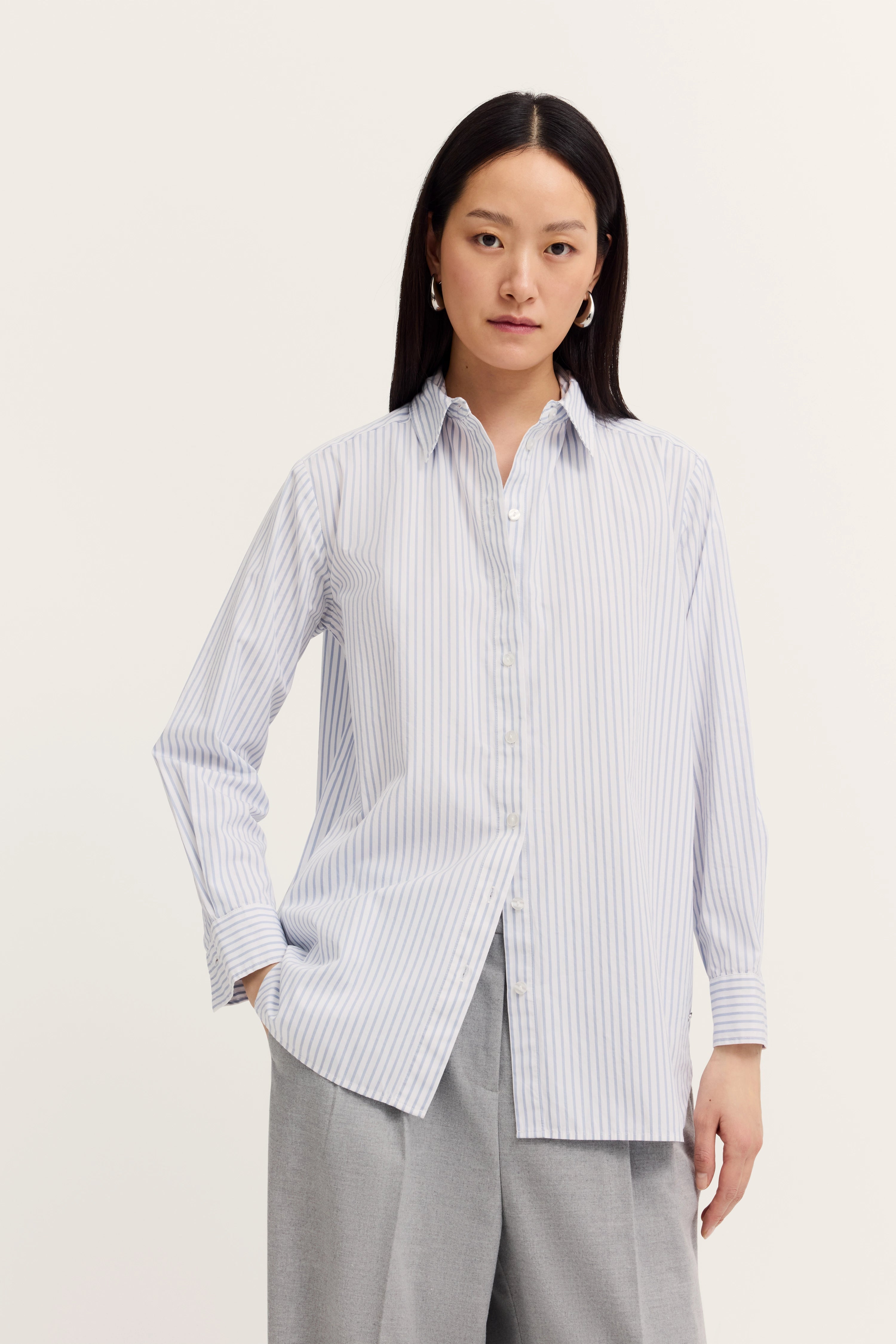 Streifenbluse in Weiß-Blau, locker getragen, kombiniert mit grauer Hose, Modelbild, entspannte Pose.