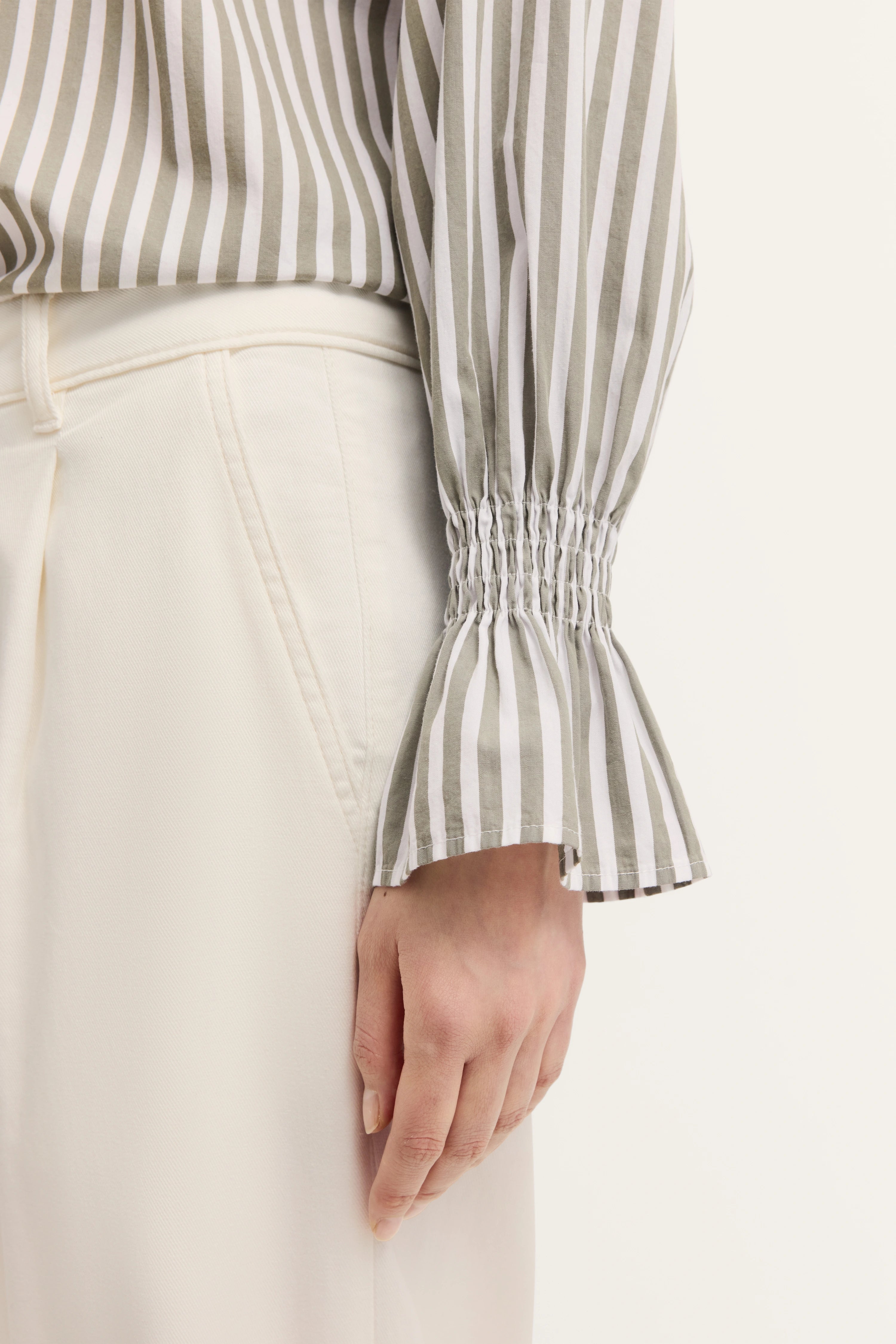 Gestreifte Bluse in Beige-Weiß mit gesmoktem Ärmel, kombiniert mit cremefarbener Hose. Büstenbild.