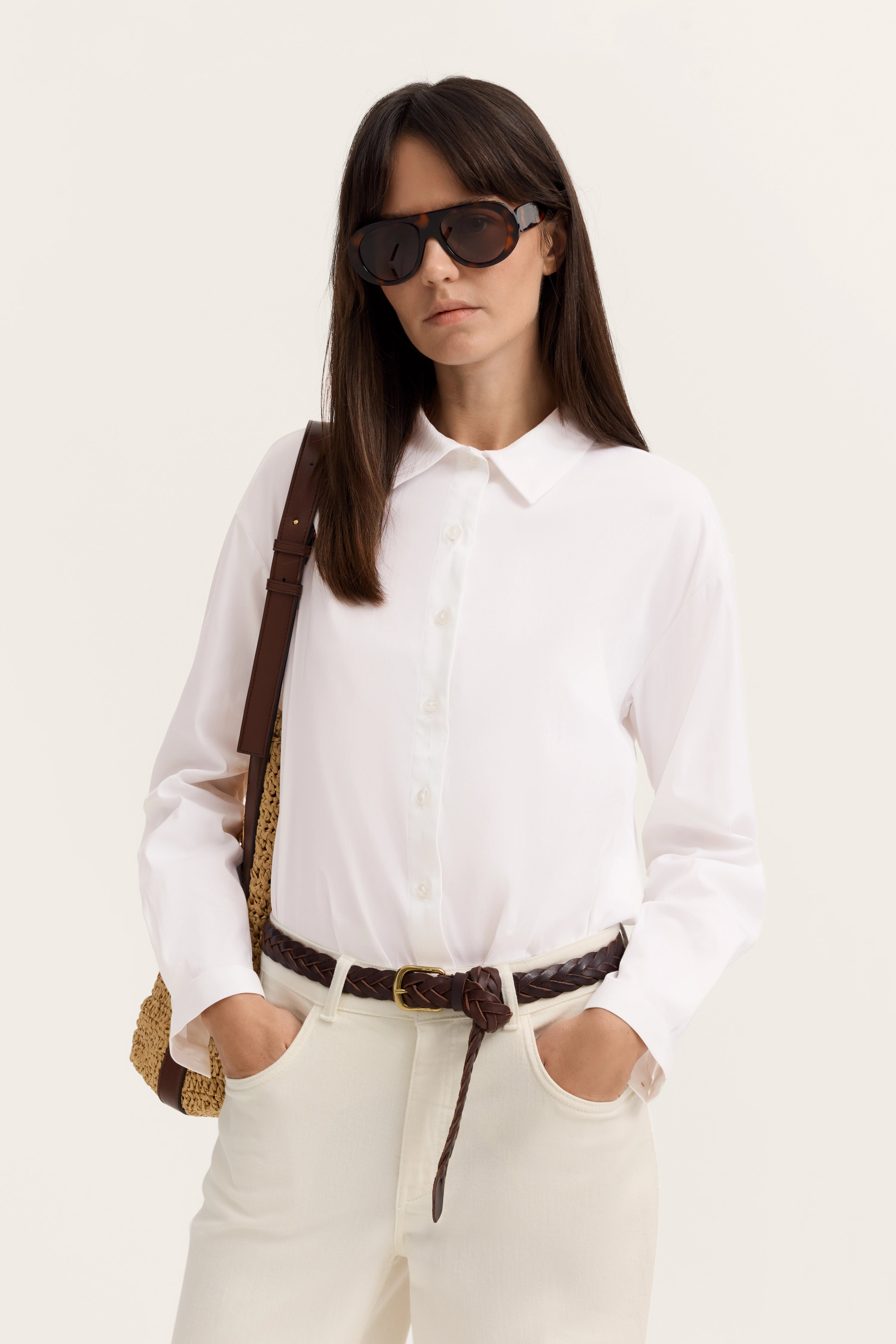Weiße Bluse, beige Hose, brauner Gürtel, Model mit Sonnenbrille, Hände in den Hosentaschen.