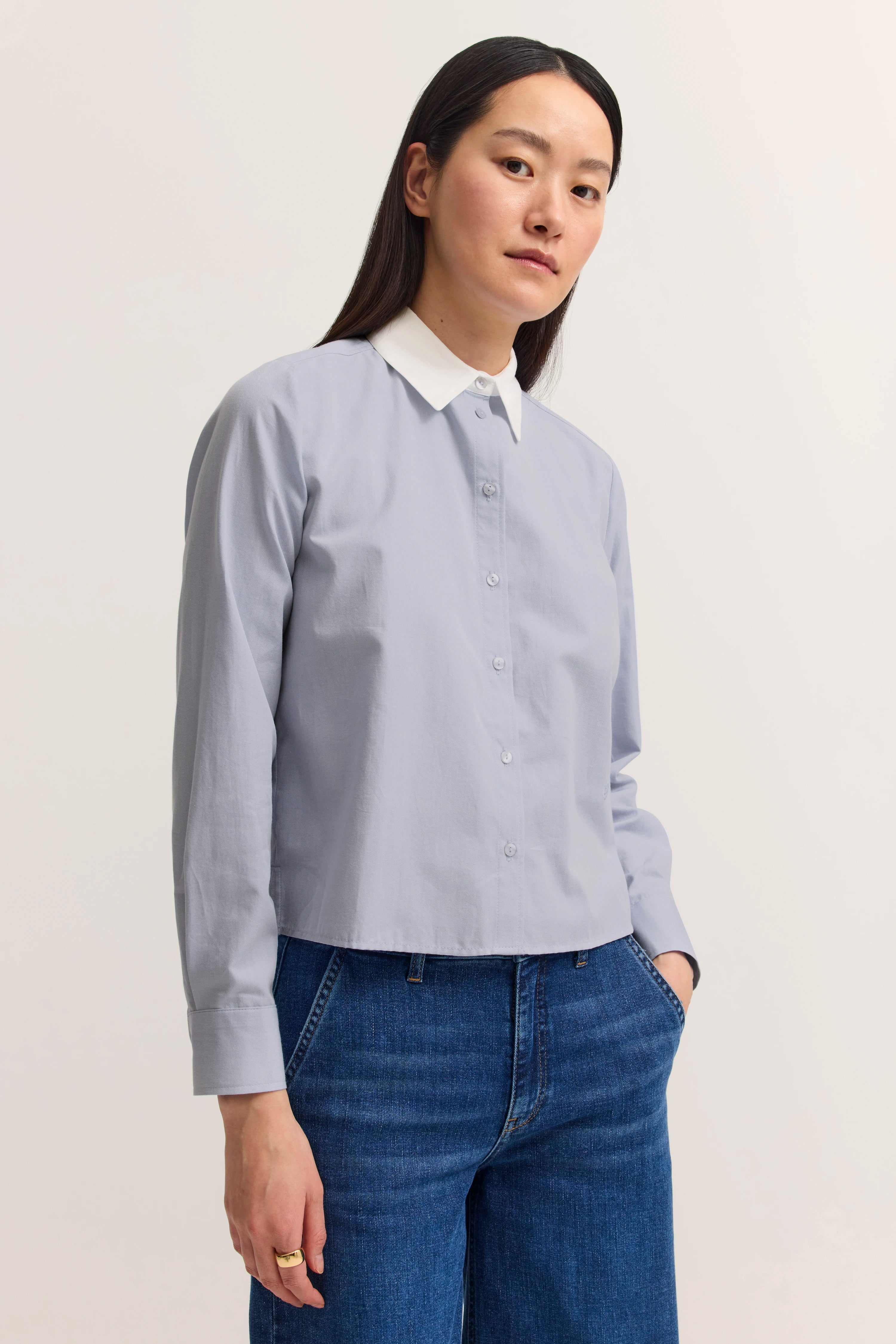 Model trägt hellblaue Bluse mit weißem Kragen, kombiniert mit blauen Jeans, Hände in den Taschen.