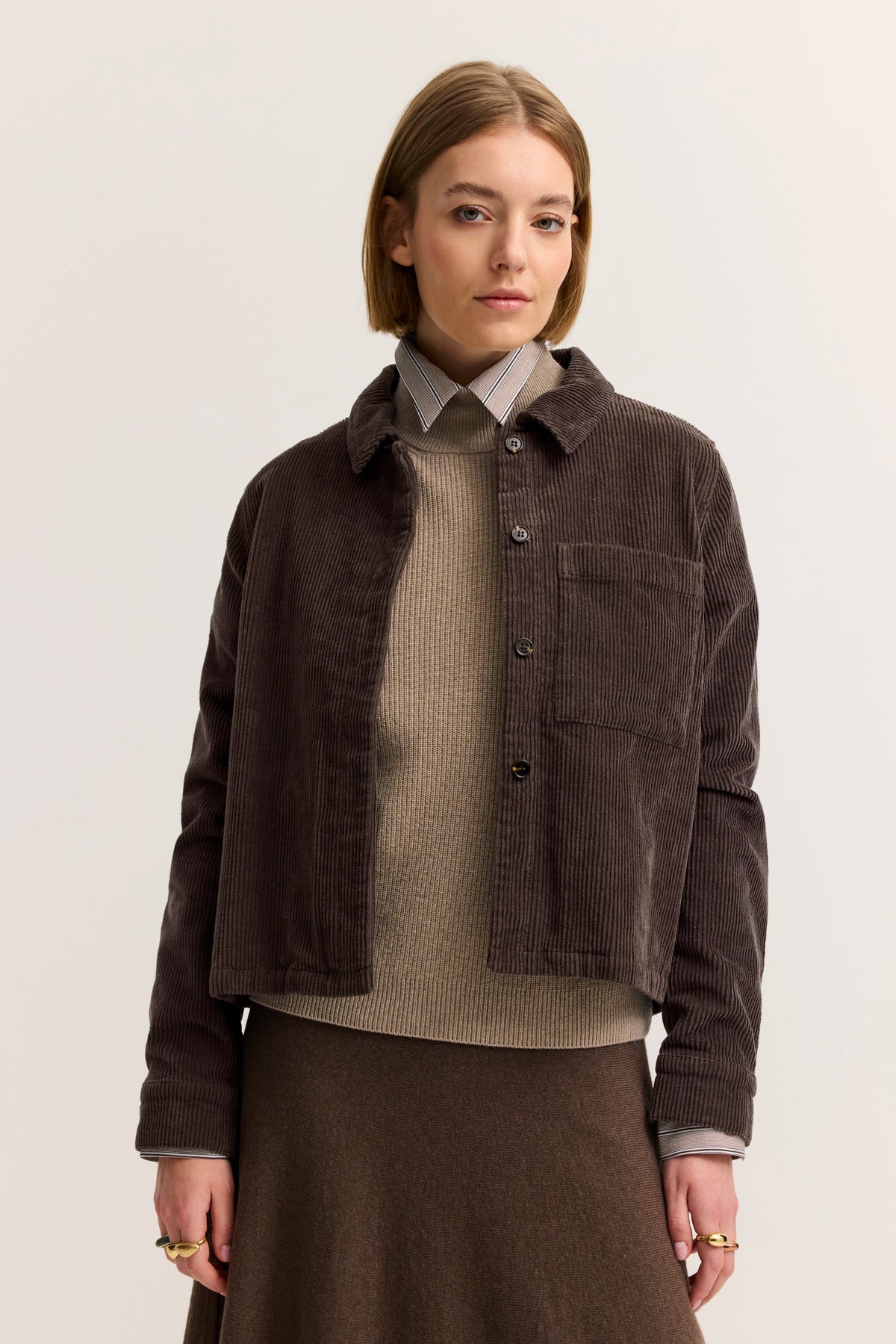 Büstenbild mit brauner Cordjacke, beigem Pullover, gestreiftem Hemd, braunem Rock. Elegantes, schlichtes Styling.