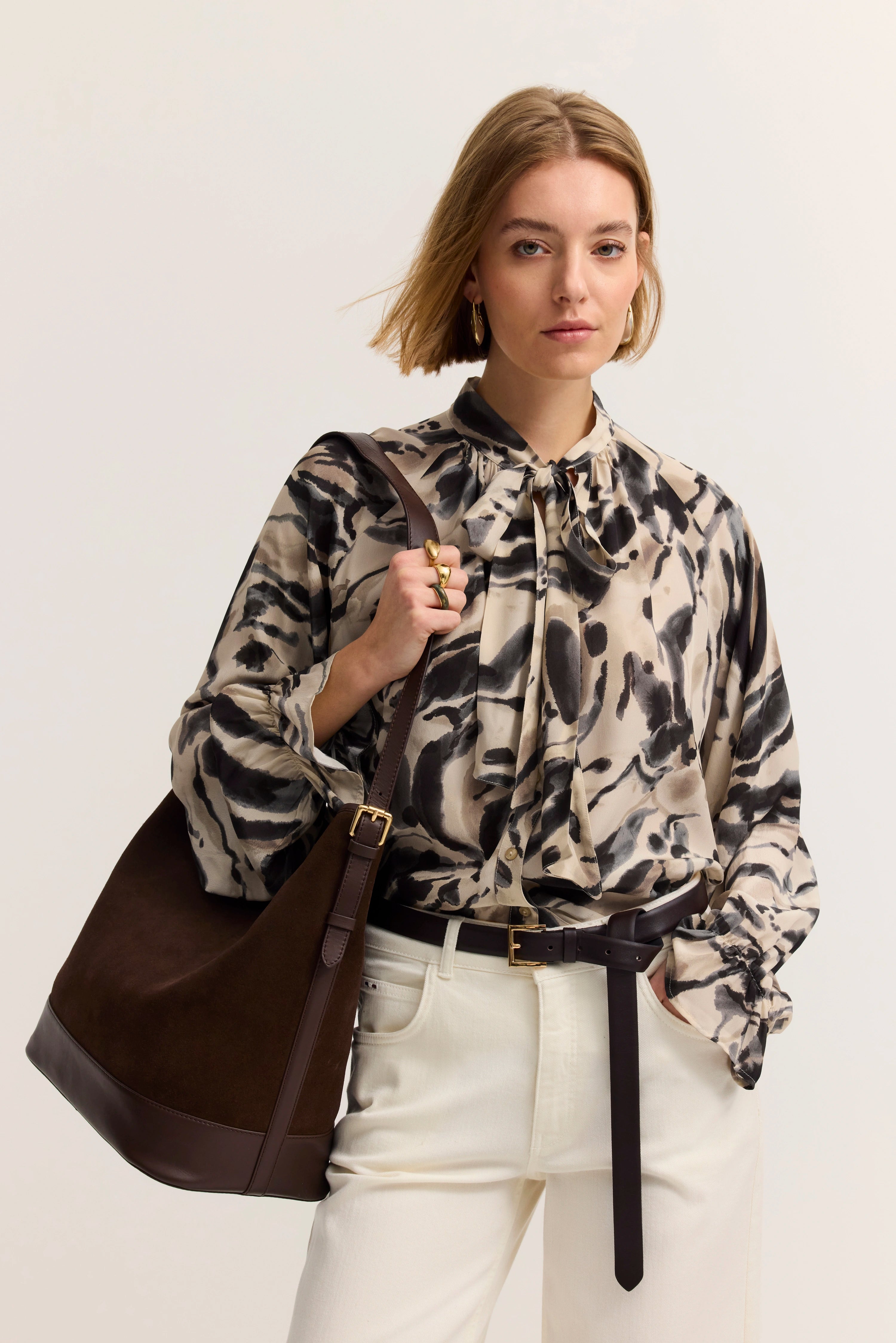 Elegantes Modelbild: Frau in gemusterter Bluse und weißer Hose, braune Tasche, selbstbewusstes Posing.