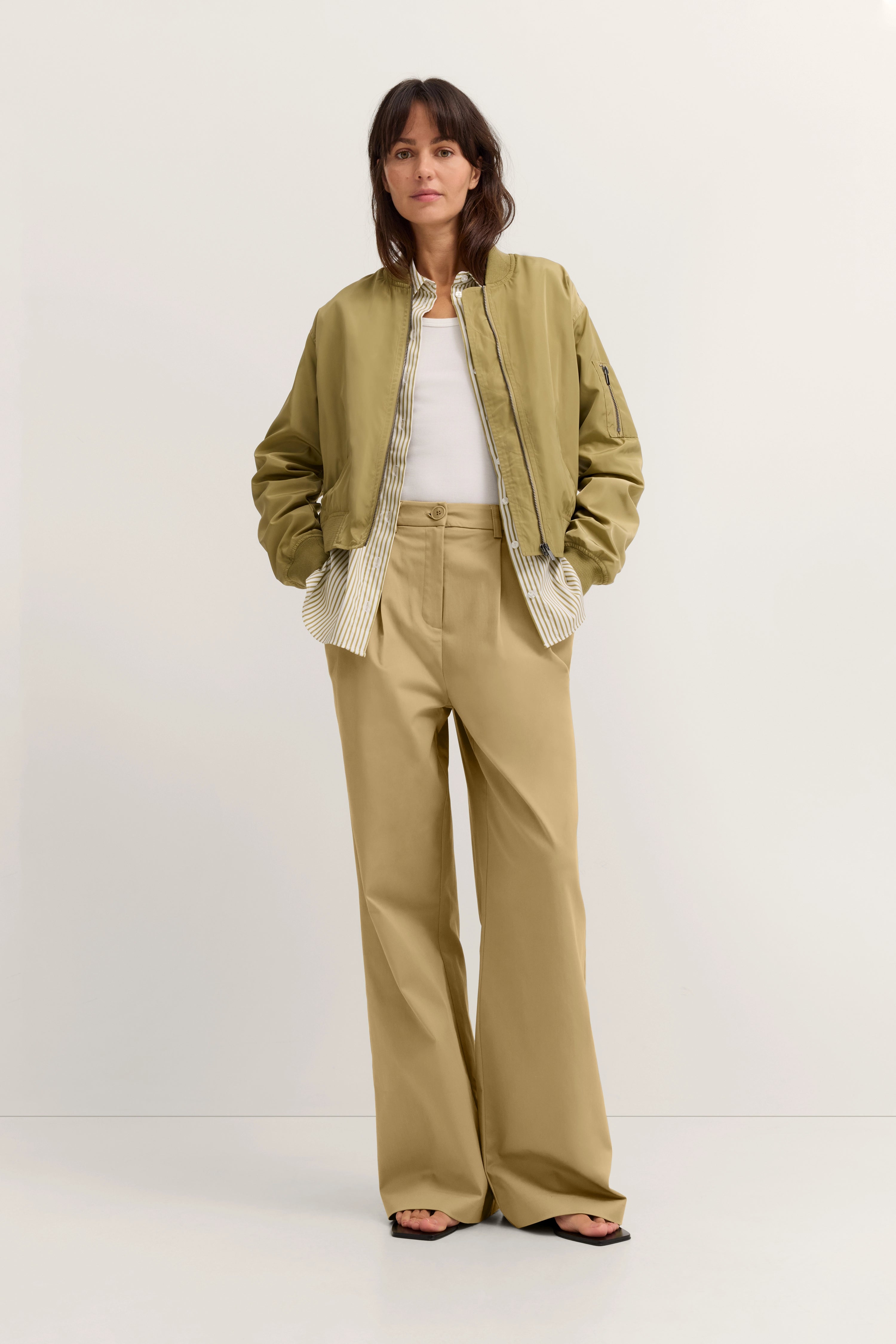 Khaki Bomberjacke, weiße Bluse, beige Hose, legeres Model, Hände in Taschen, bugatti Stil.