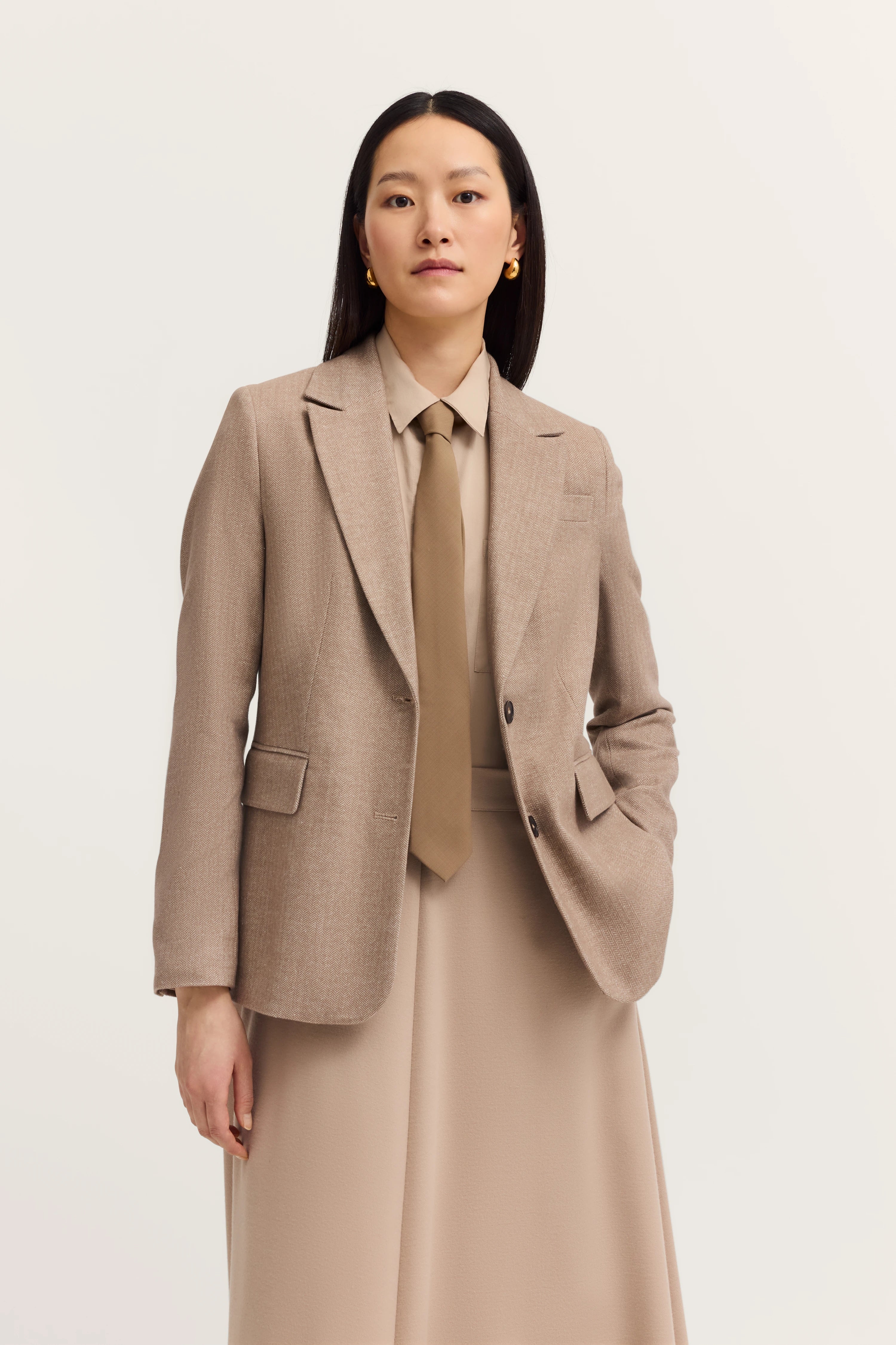 Model in beige blazer and skirt, beige shirt and tie, posing with hand in pocket.