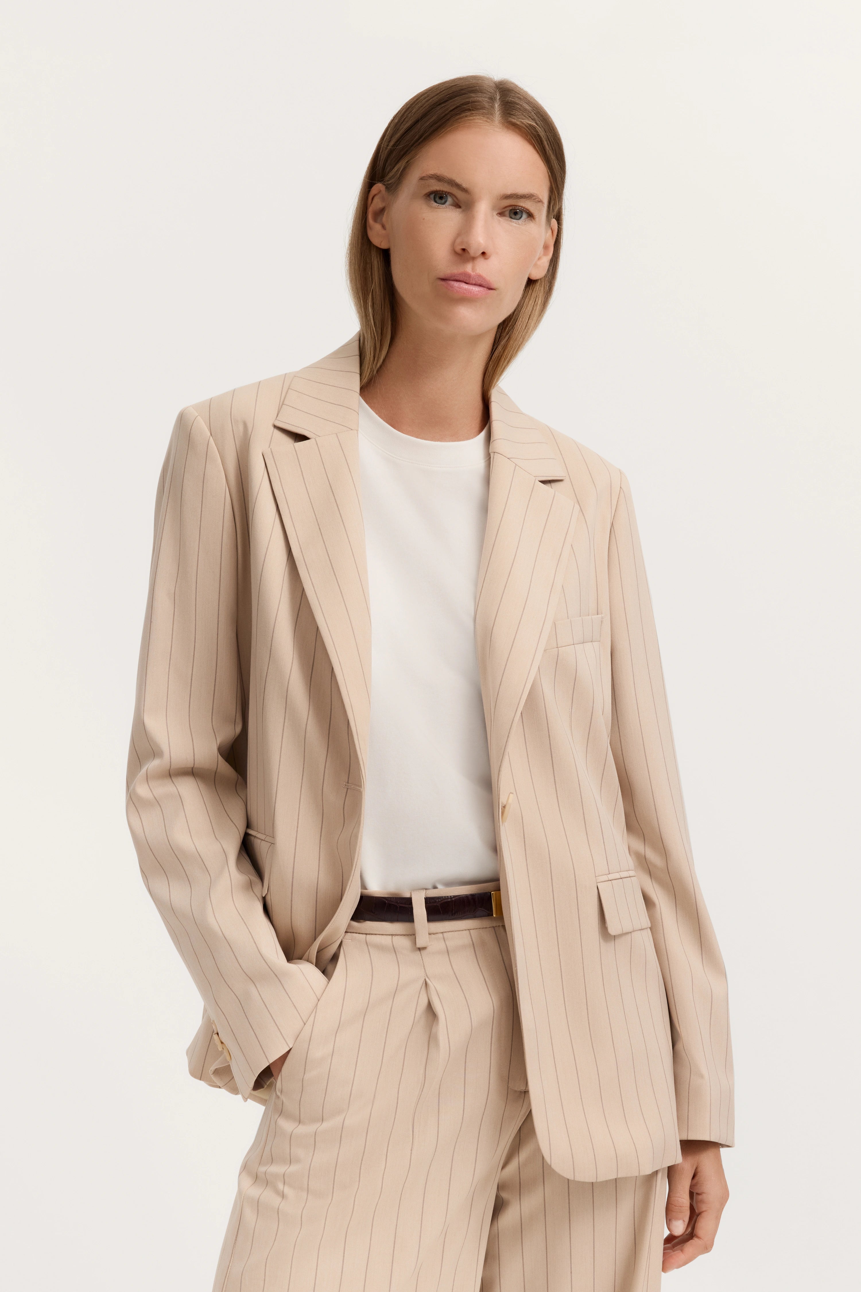 Beiges Nadelstreifen-Outfit mit Blazer und Hose, weißes Shirt, Modelpose mit Hand in der Tasche.