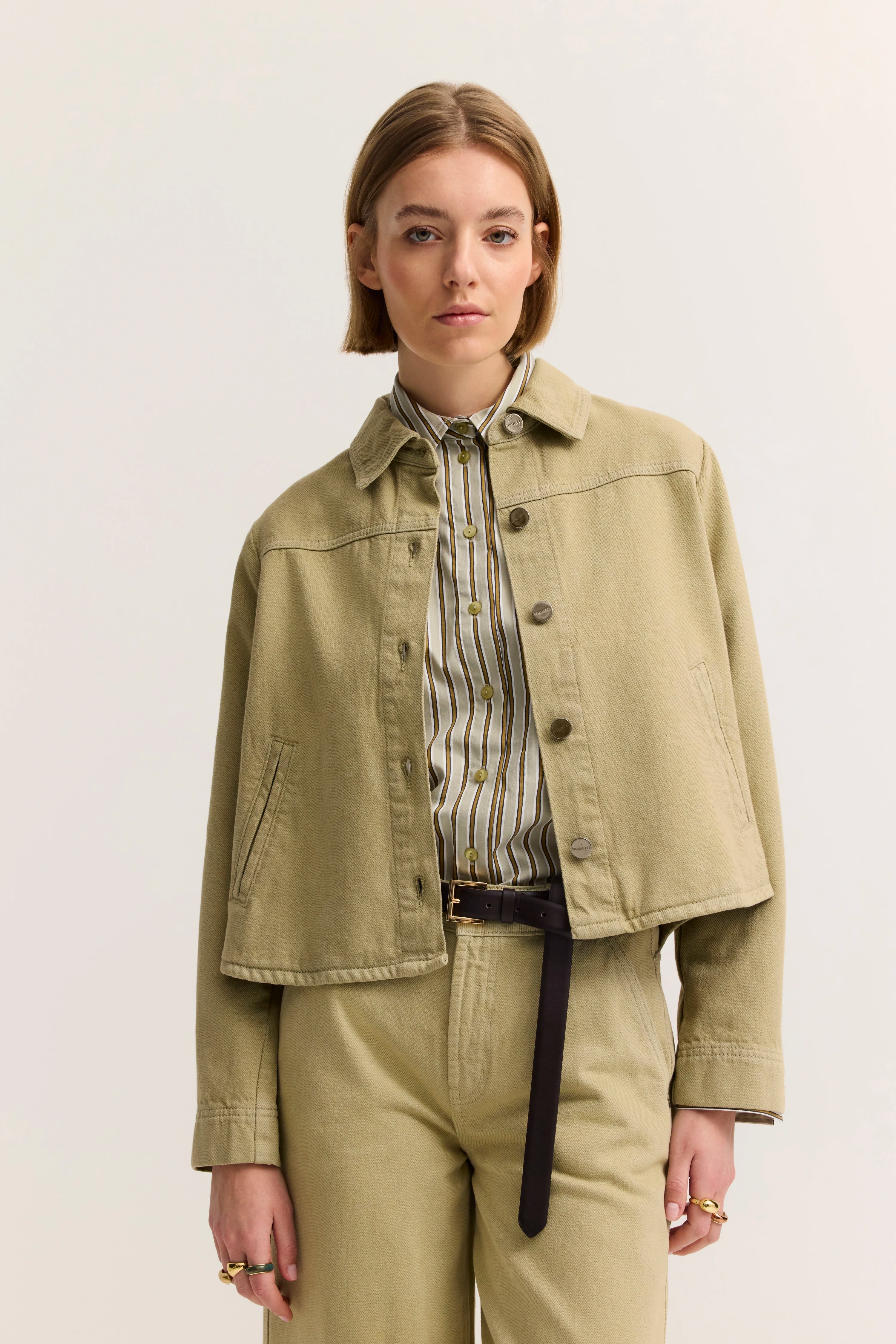 Model trägt beigefarbene Jacke und Hose, gestreiftes Hemd. Posenbild, lockere Haltung.