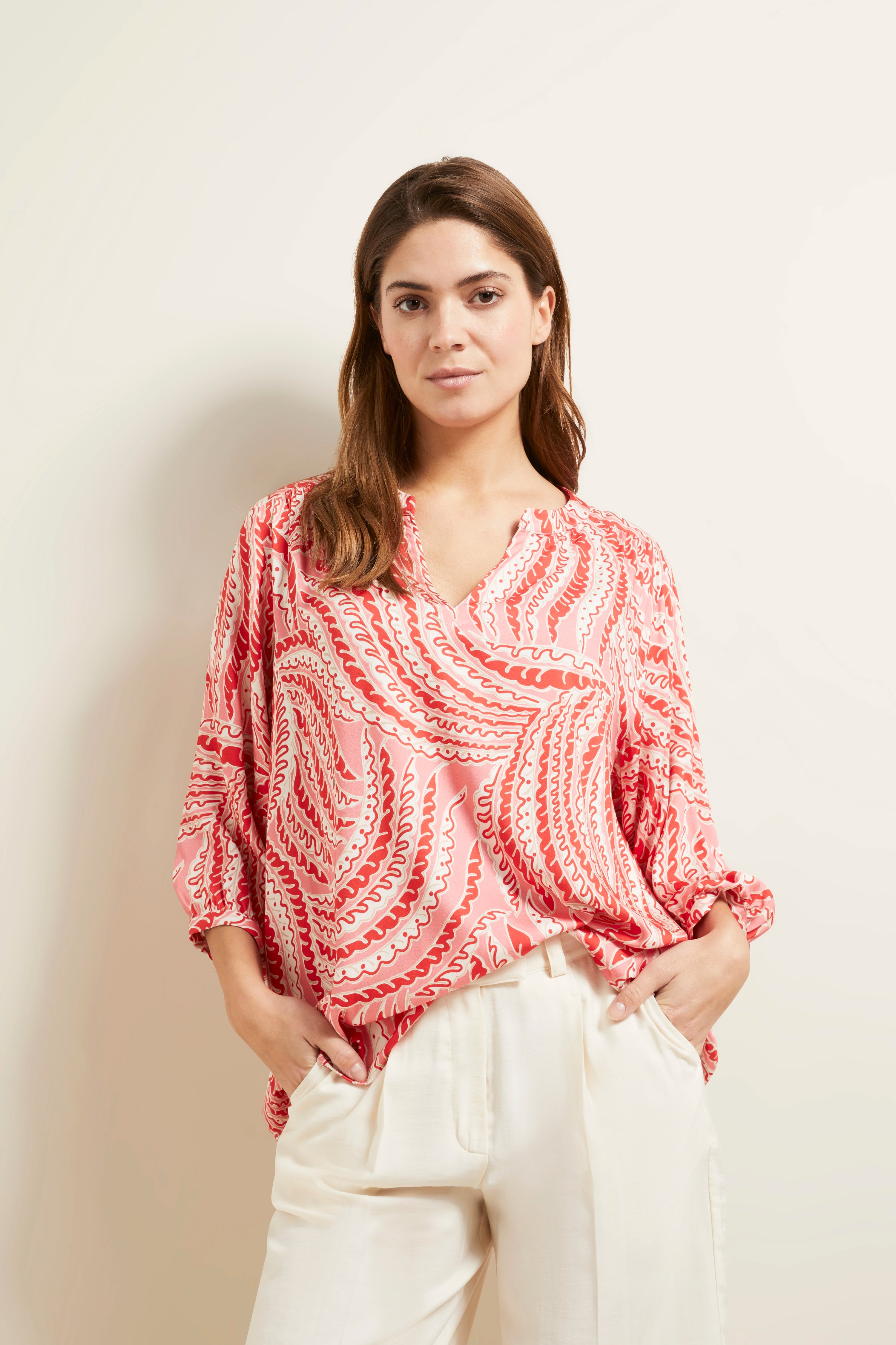 Rosa gemusterte Bluse, weiße Hose, Model stehend mit Händen in den Taschen, bugatti Stil.