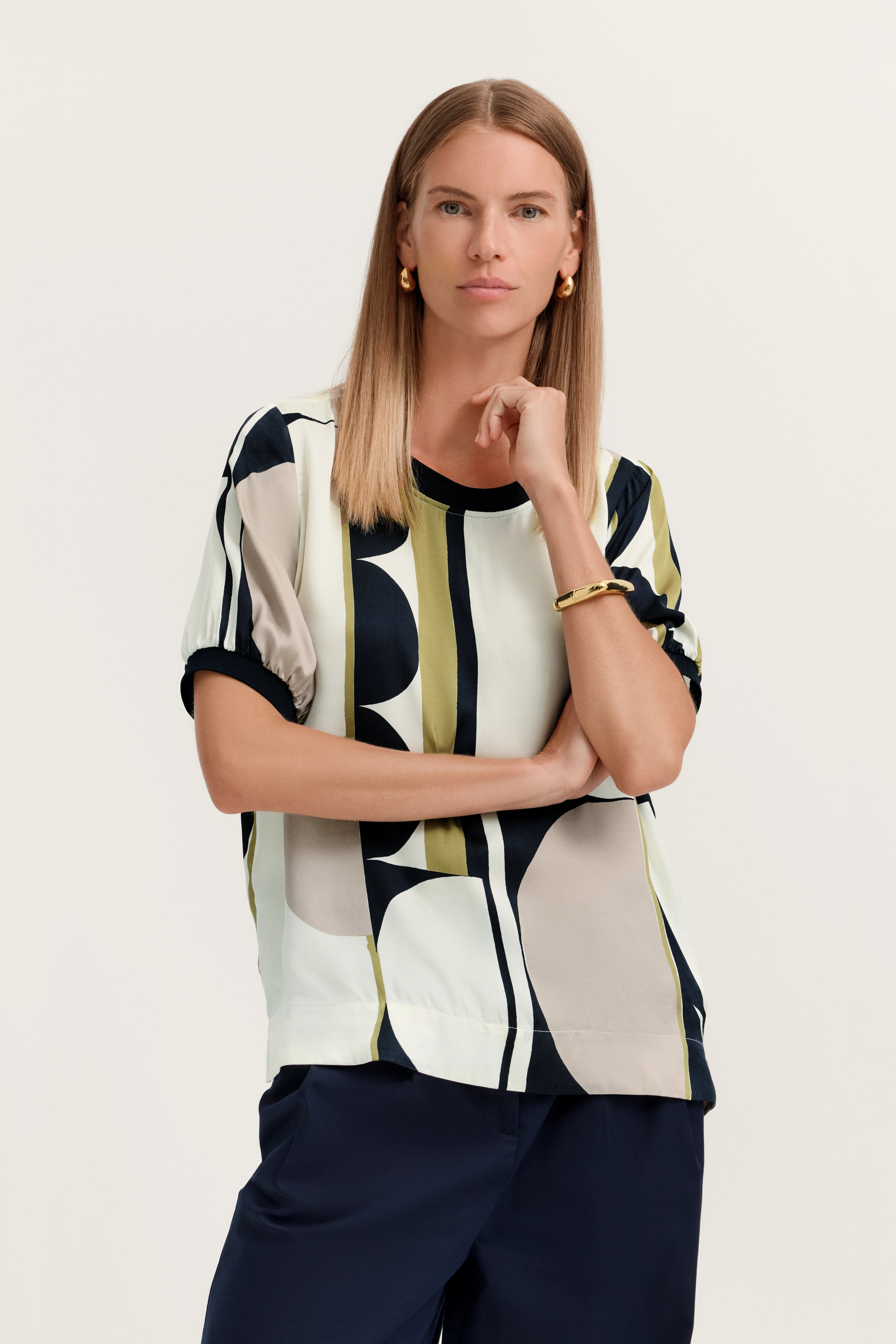 Model in gemustertem bugatti-Shirt, Halbarm, und blauer Hose. Pose: Stehen mit Hand am Kinn.