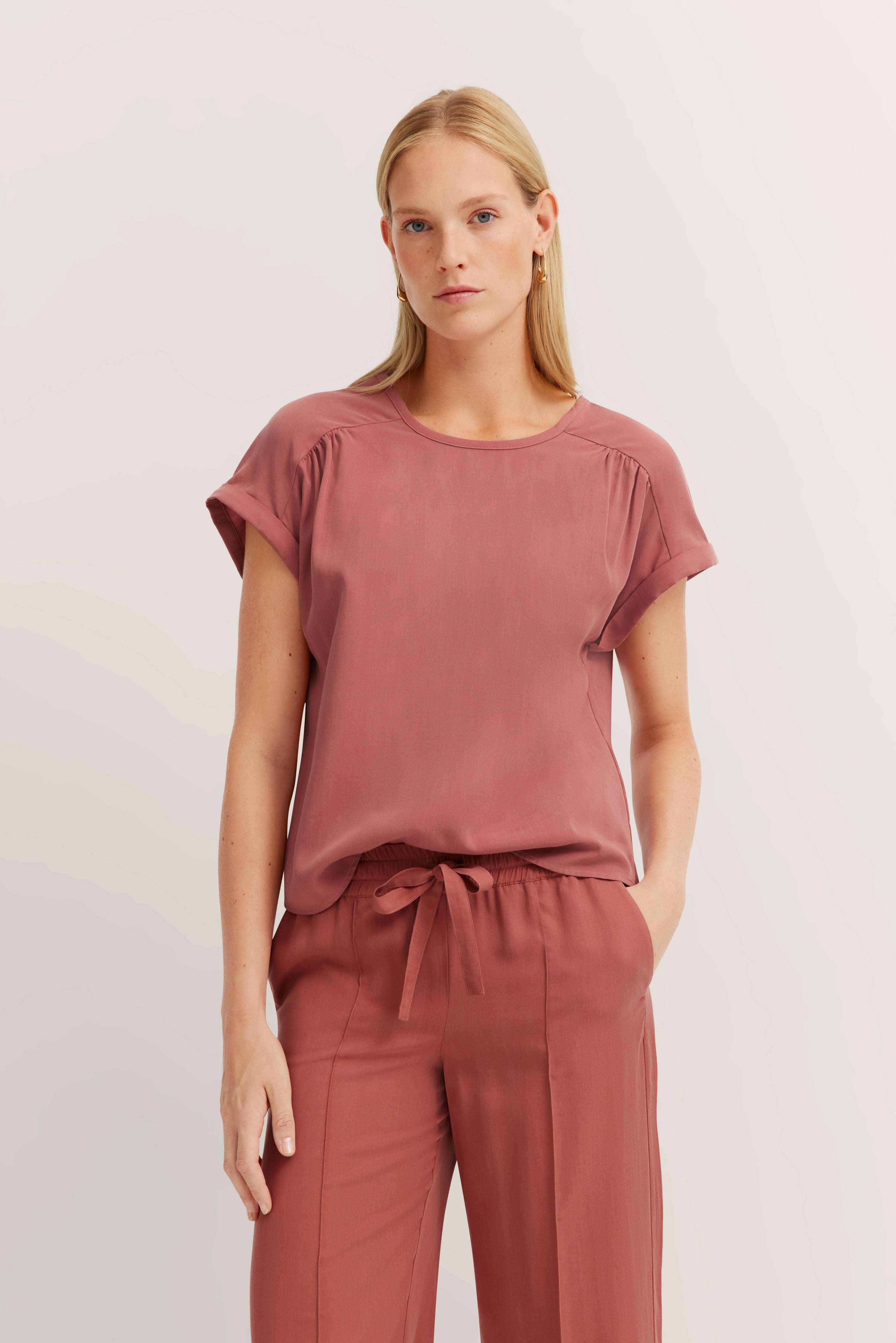 Model in rosékleurige bugatti outfit, los T-shirt en bijpassende broek, ontspannen pose.