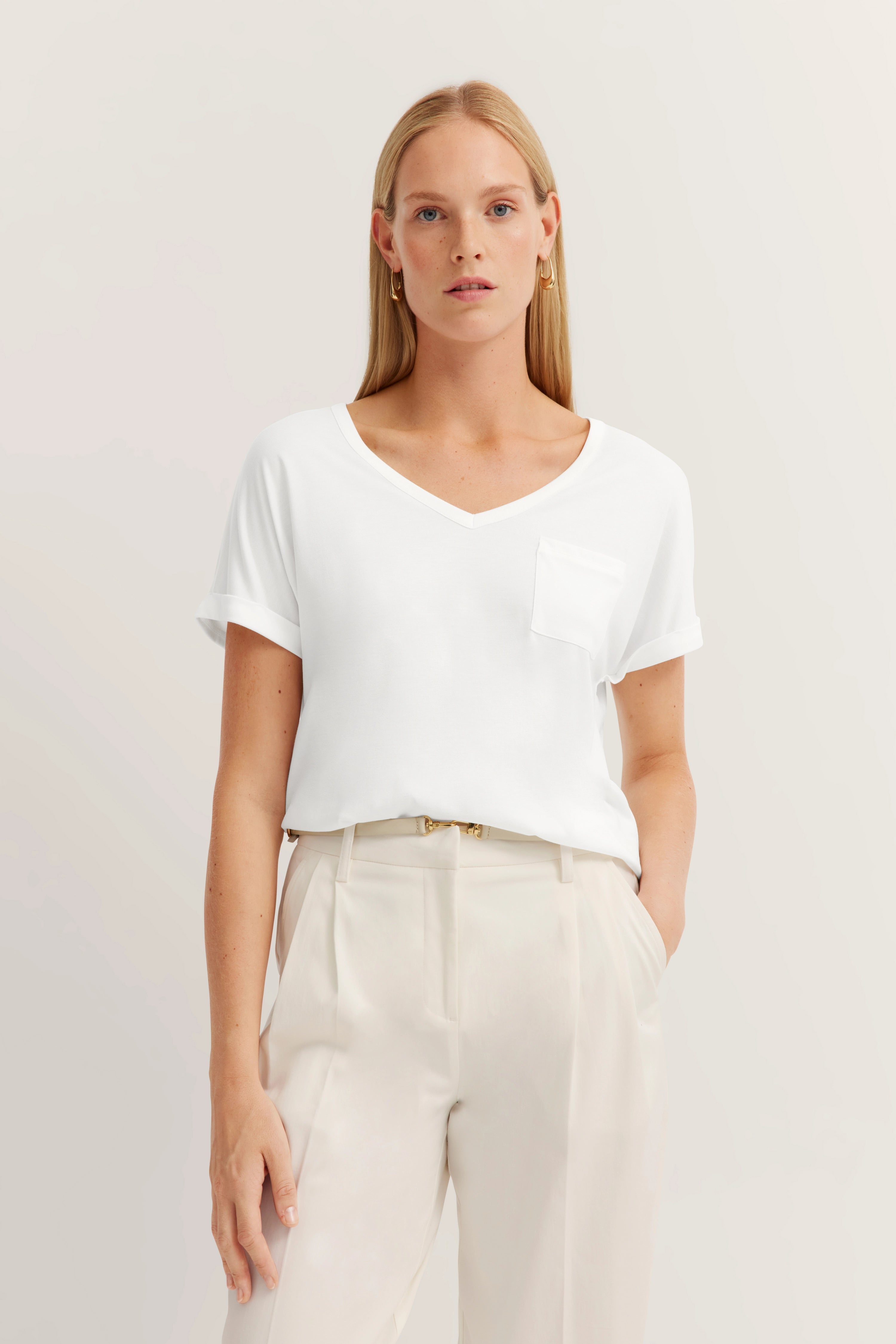 Weiße Bluse mit V-Ausschnitt, cremefarbene Hose, Model in entspannter Pose.