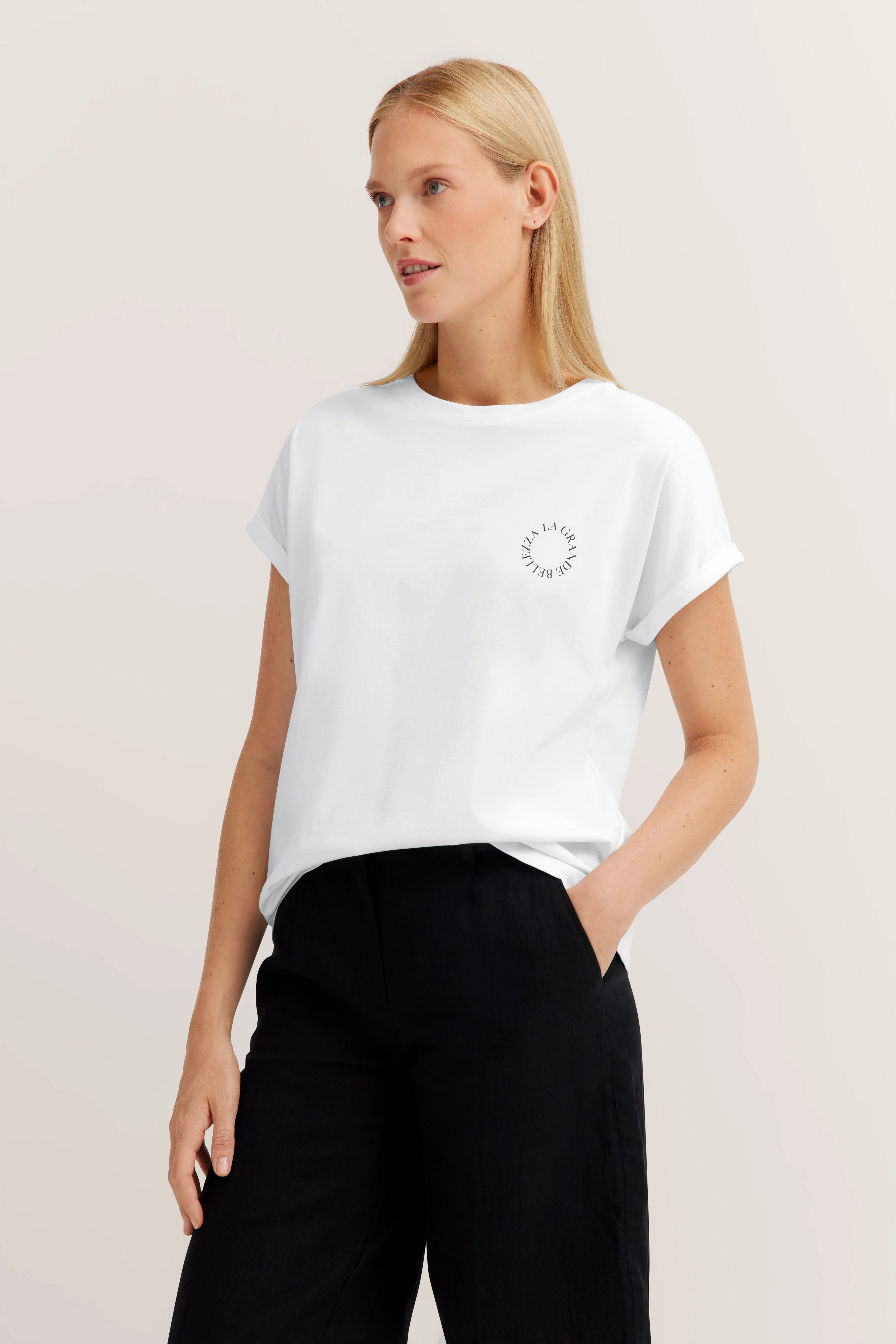 Weißes T-Shirt mit Aufdruck, schwarze Hose, legeres Modelbild, entspannte Pose.