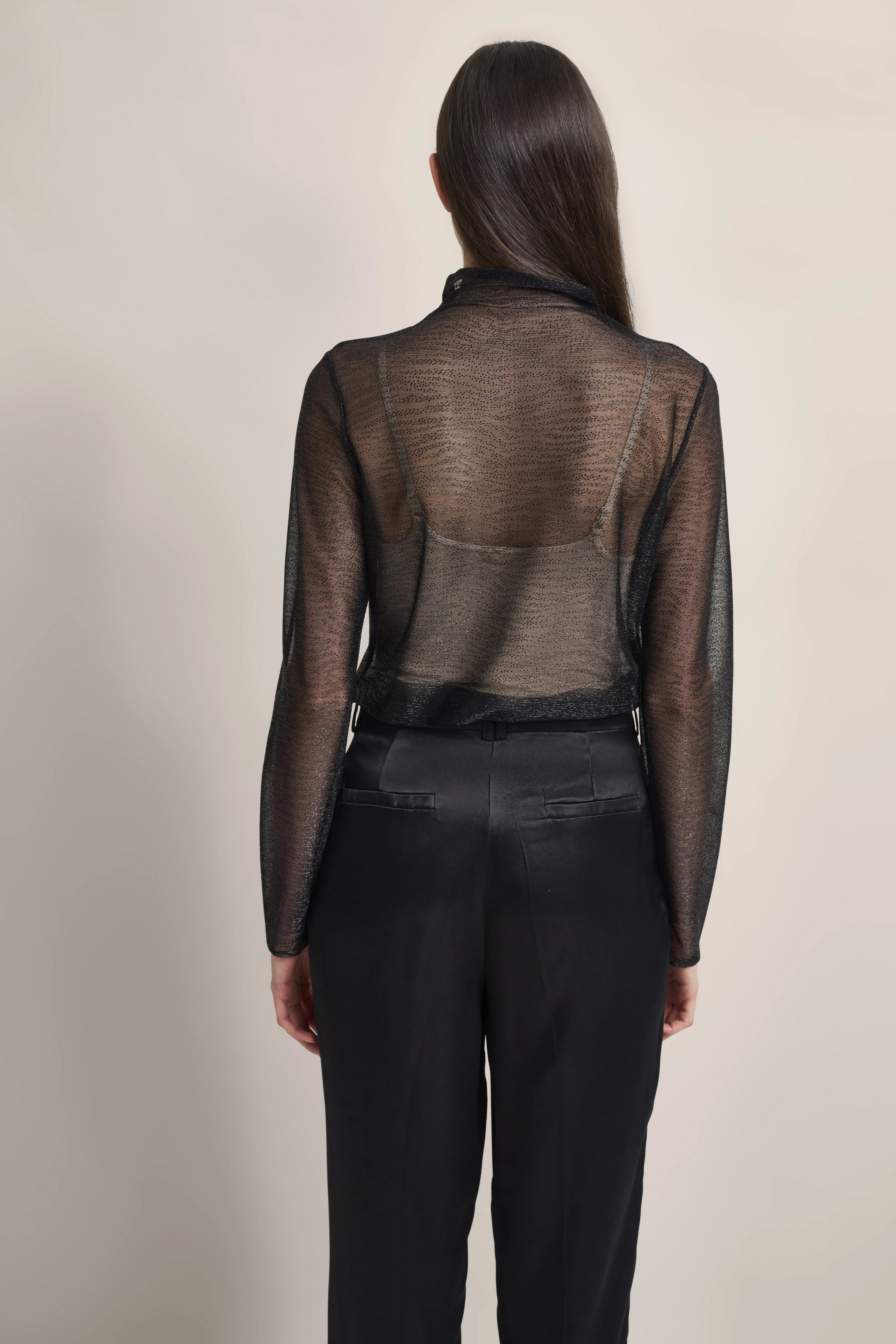 Black sheer blouse with black pants, model from behind, casual stance.