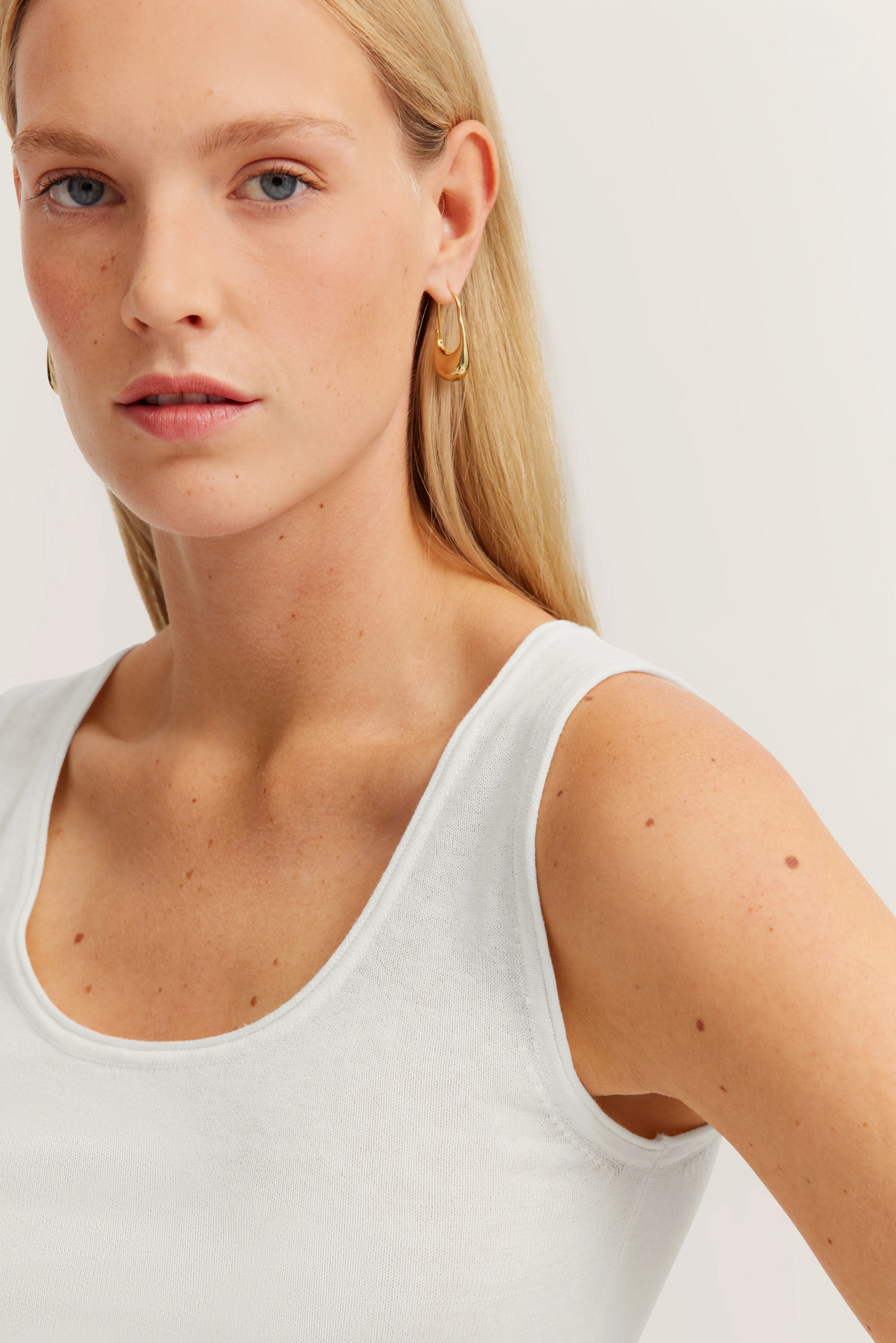 Model in wit mouwloos top, gouden oorbel, ontspannen pose.
