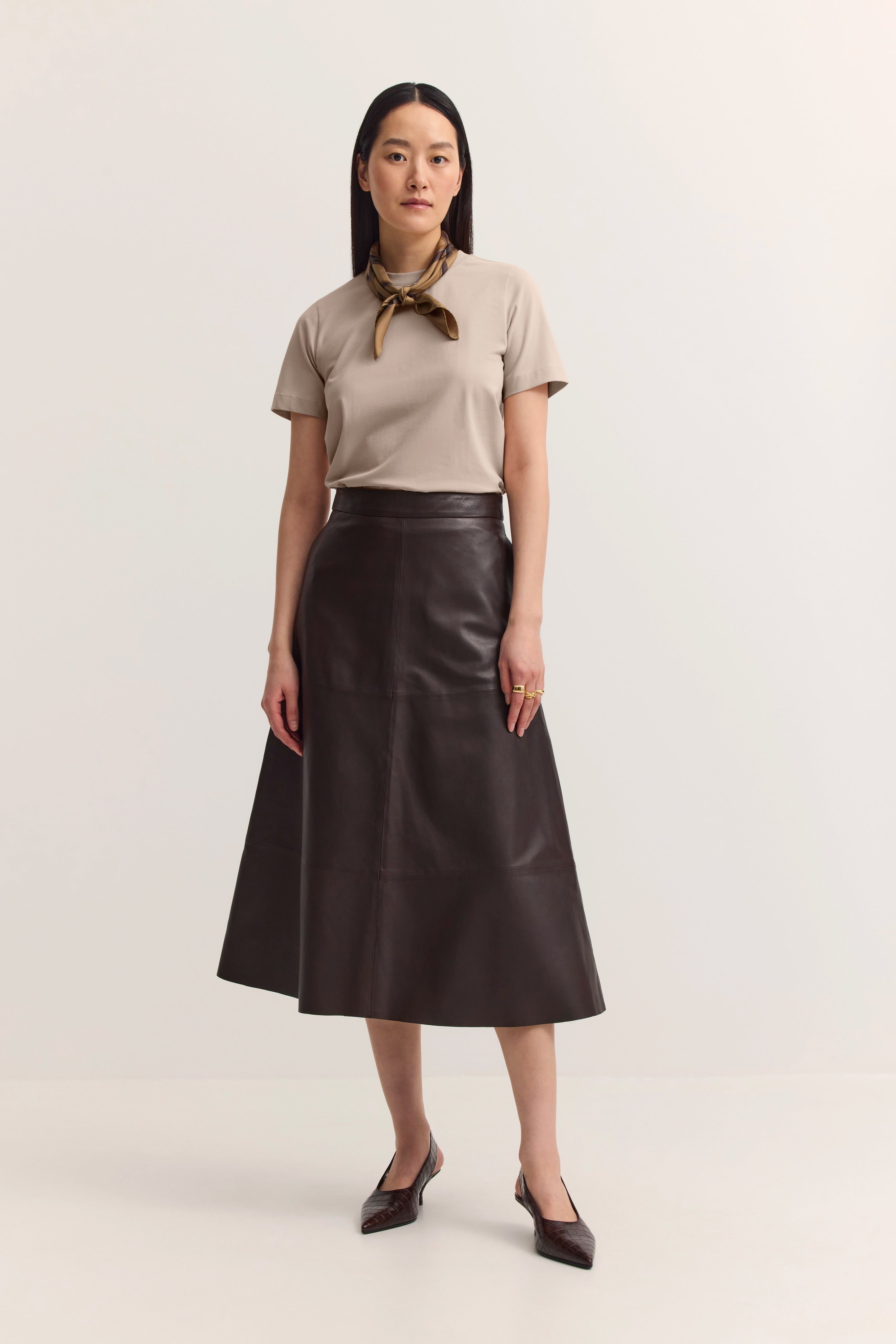Model in beigem Shirt, braunem Lederrock und Halstuch, steht frontal, Eleganz und Stil von bugatti.