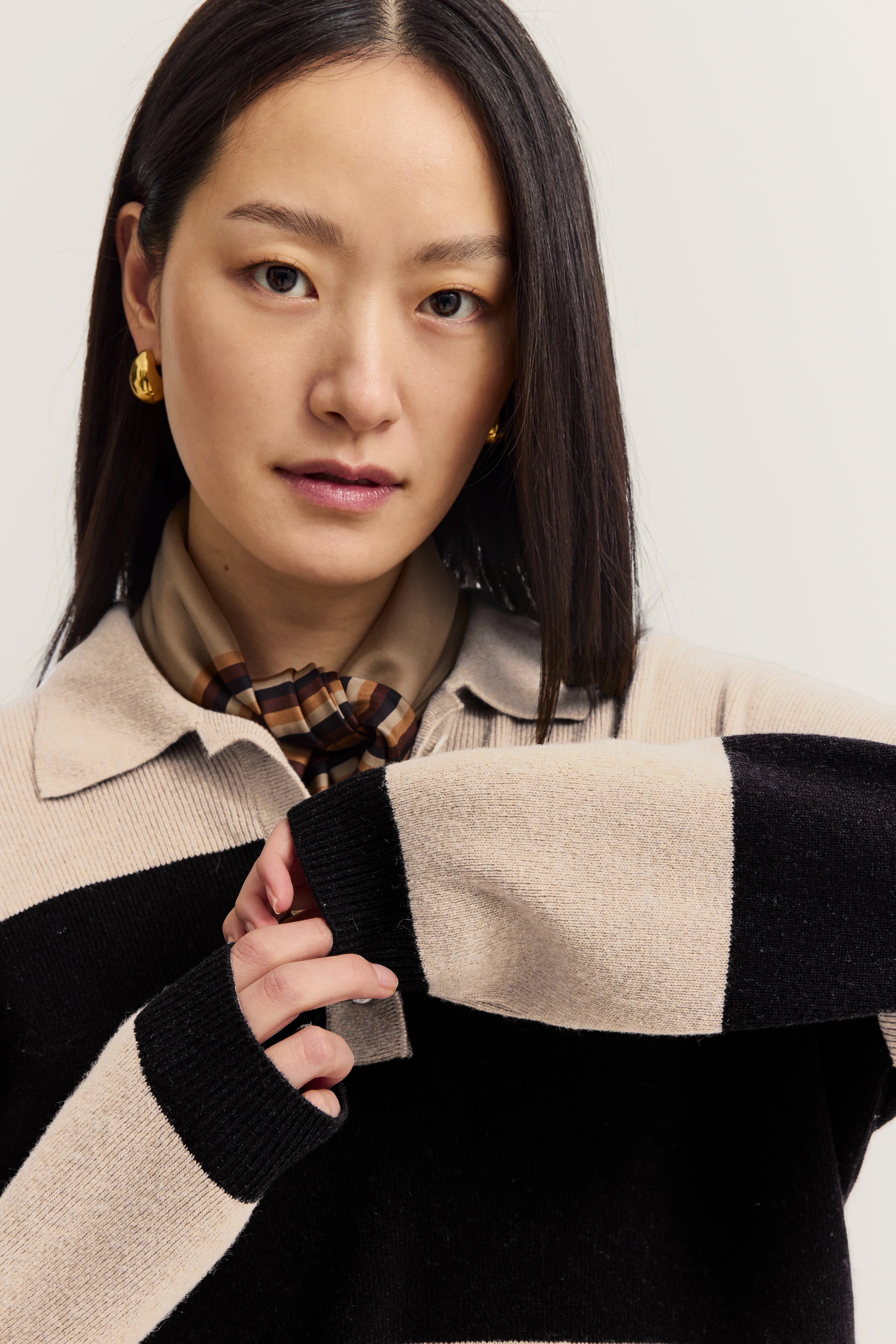 Büstenbild von Frau in gestreiftem Pullover, beige-schwarz, mit kariertem Schal und goldenen Ohrringen.