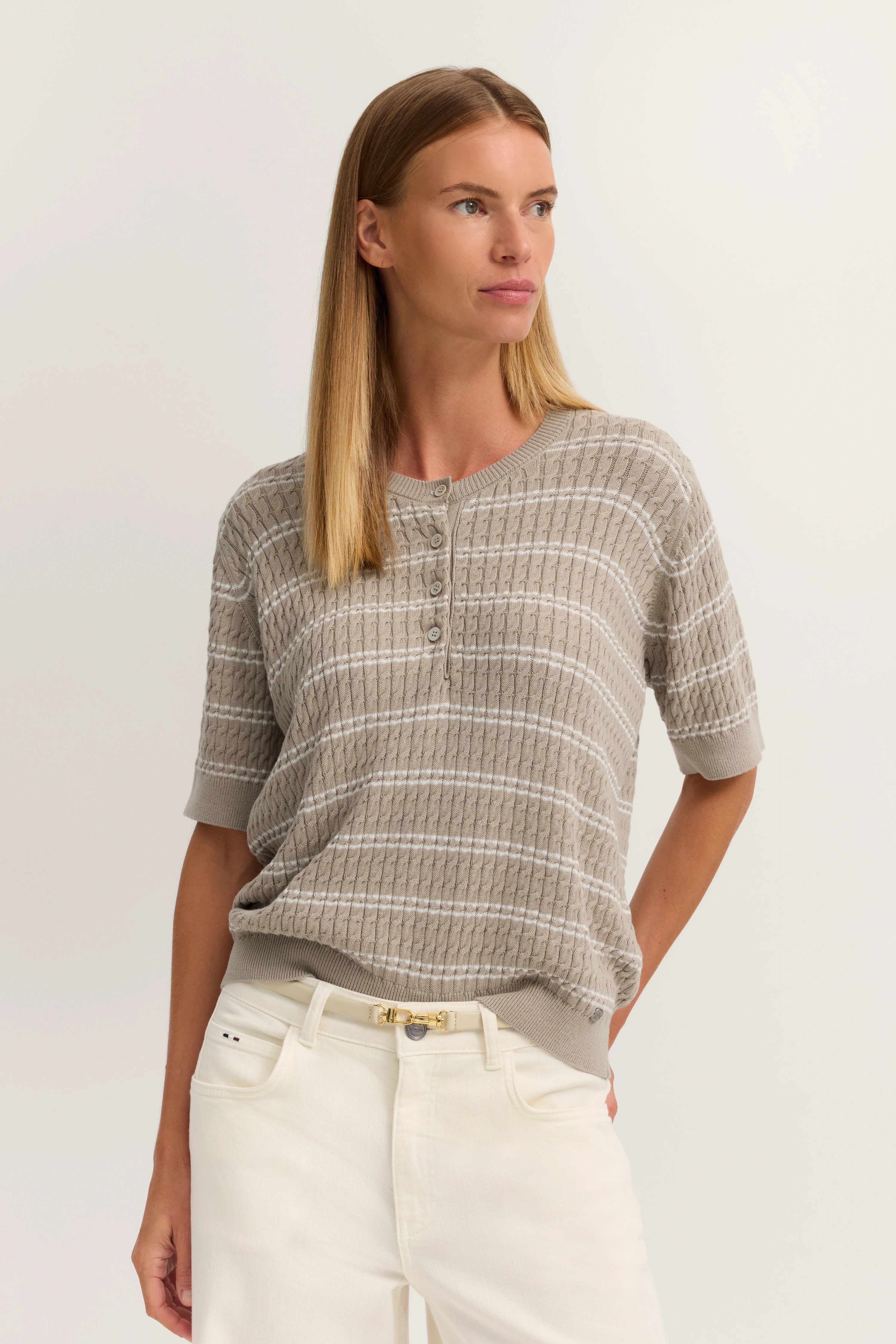 Beige Strickshirt mit Streifen und Knopfleiste, kombiniert mit weißen Hosen, Modelbild, entspannte Pose.