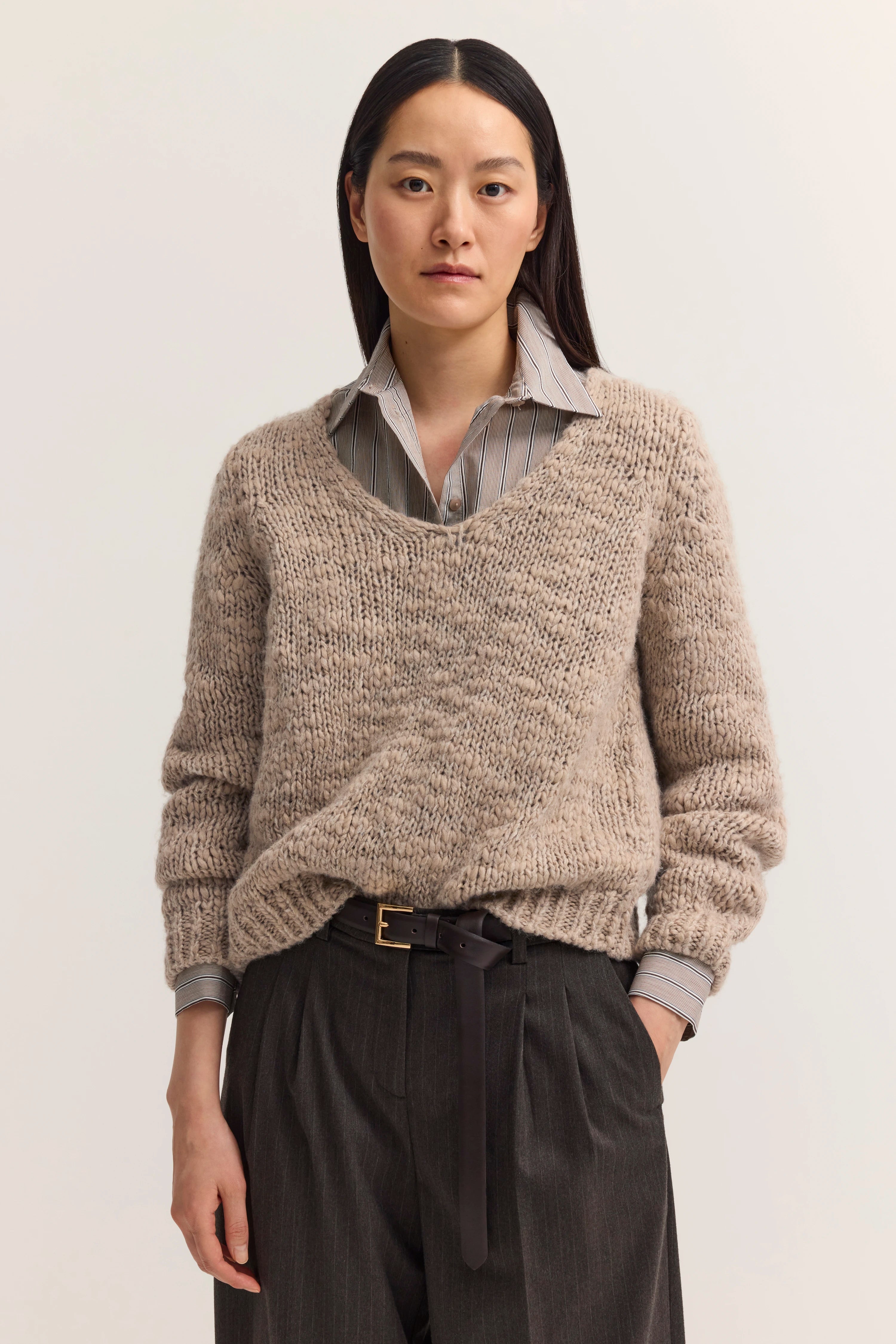 Beiges Strickpullover-Outfit mit gestreiftem Hemd und grauer Hose. Modelbild, entspanntes Posing.