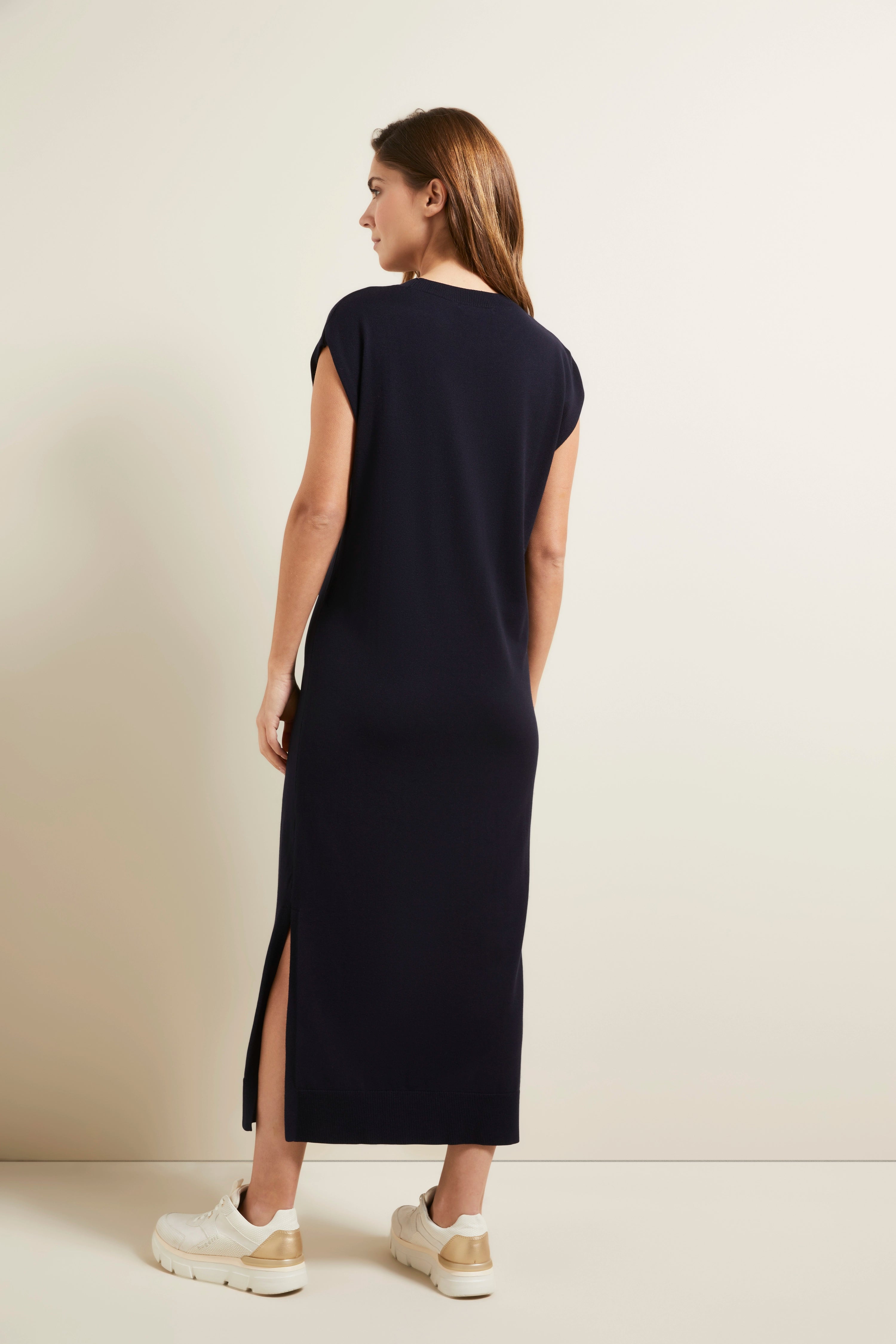 Model in langem, dunkelblauem bugatti Kleid, Rückenansicht, entspanntes Posing mit weißen Sneakern.