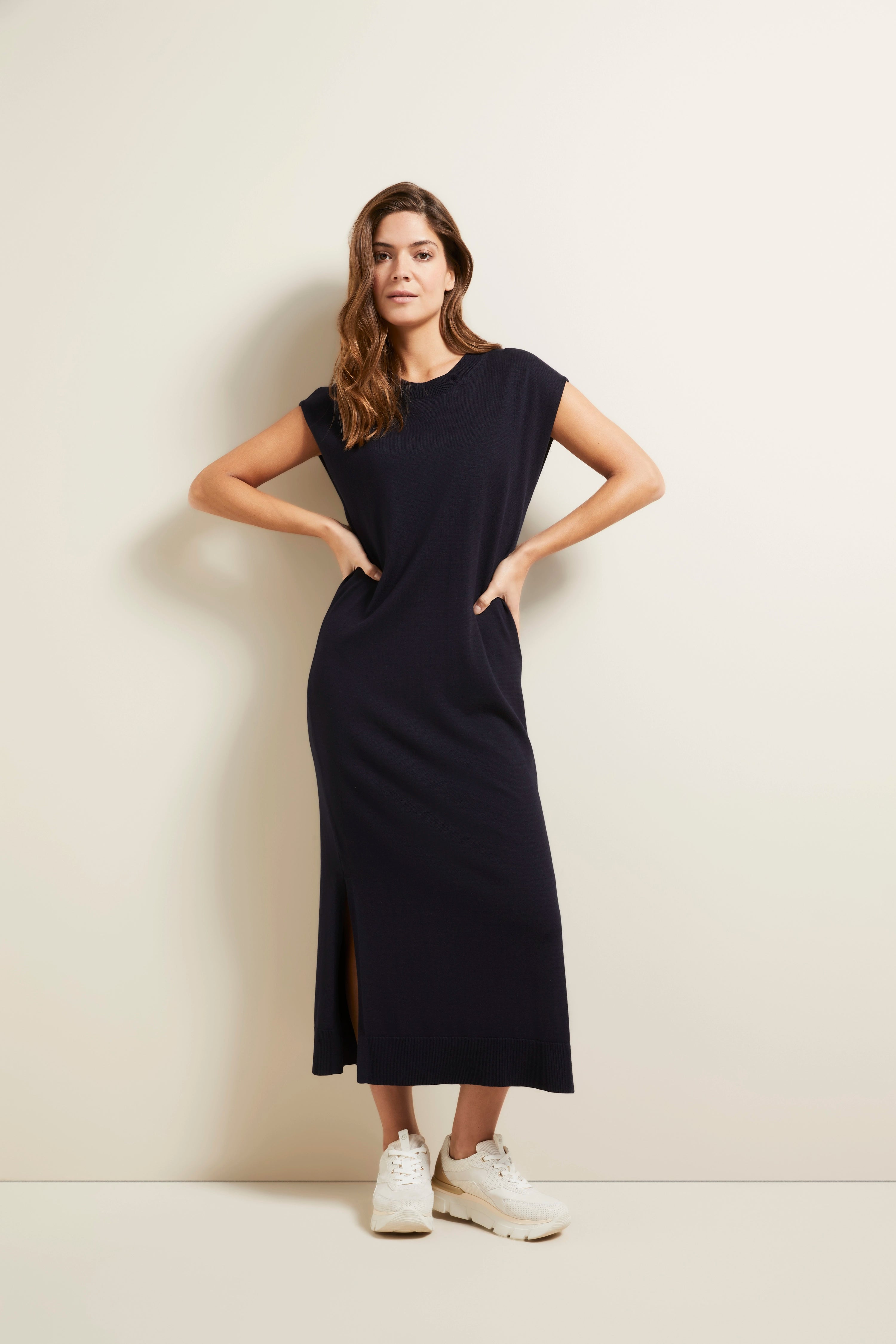 Model draagt zwarte bugatti maxi-jurk met zijsplit, witte sneakers, ontspannen pose.