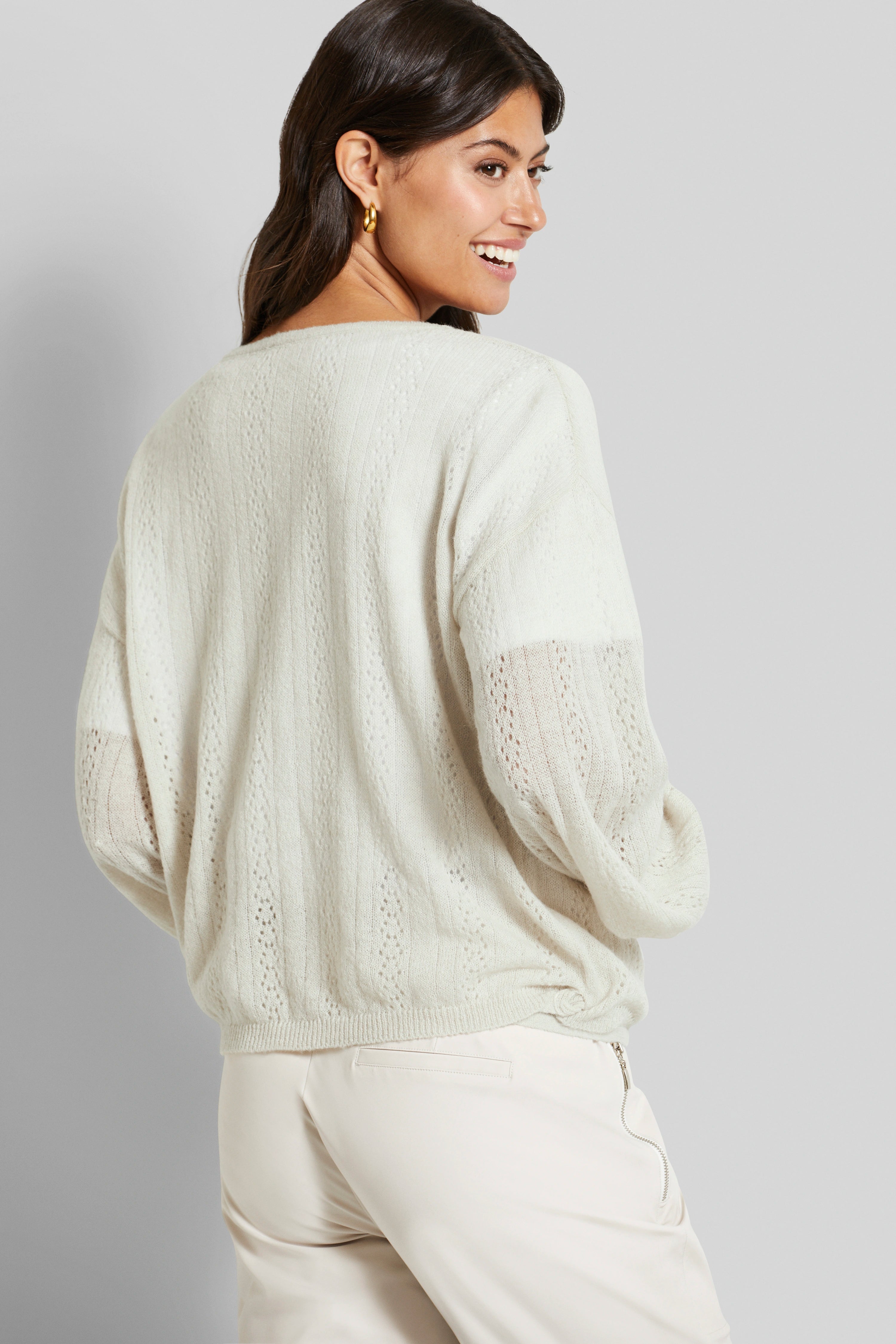 Beige gebreide trui met patroon, model lacht over schouder, lichtbeige broek, bugatti stijl.