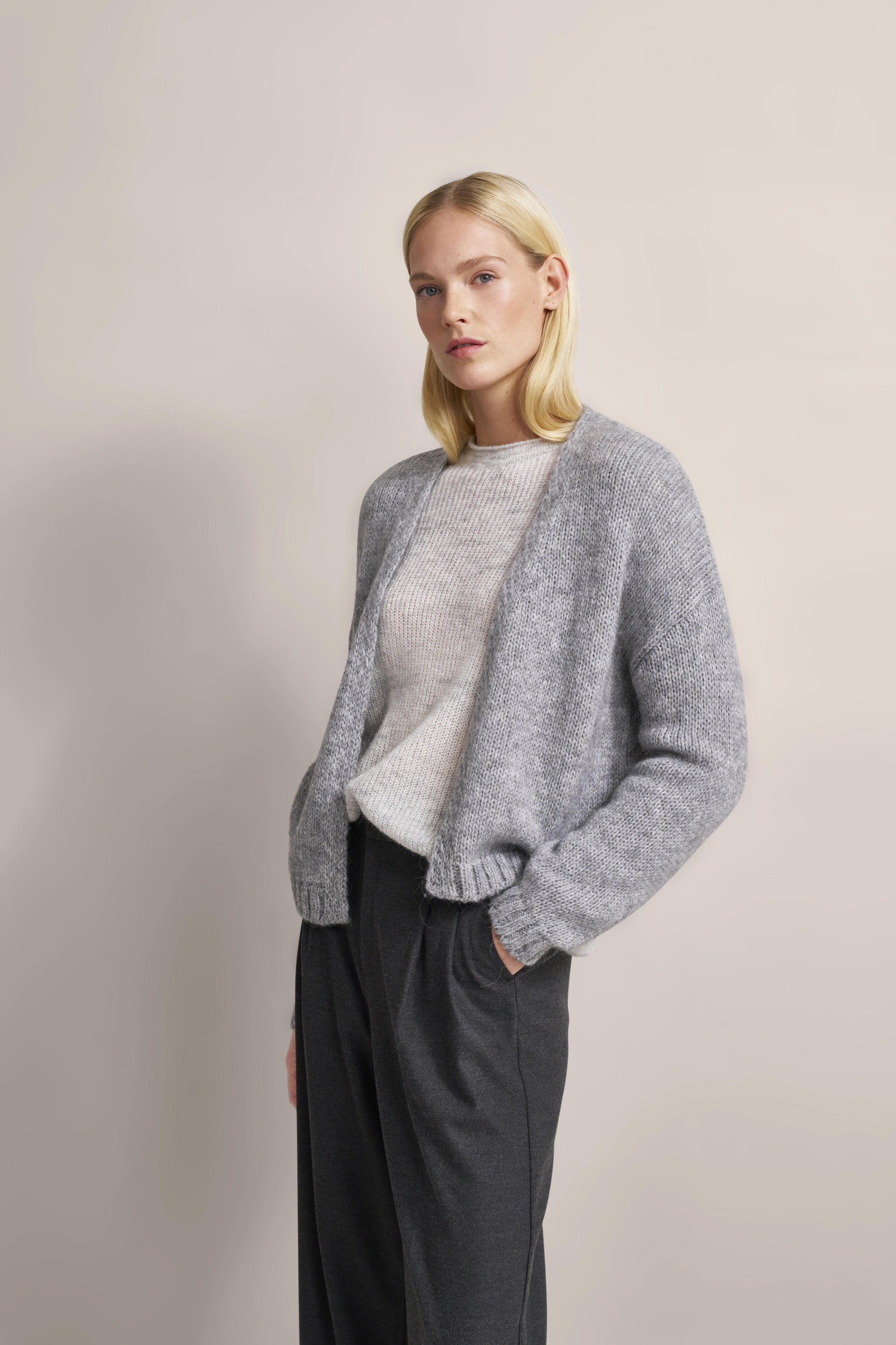 Grauer Cardigan über cremefarbenem Pullover, schwarze Hose. Modelbild, entspanntes Posing.