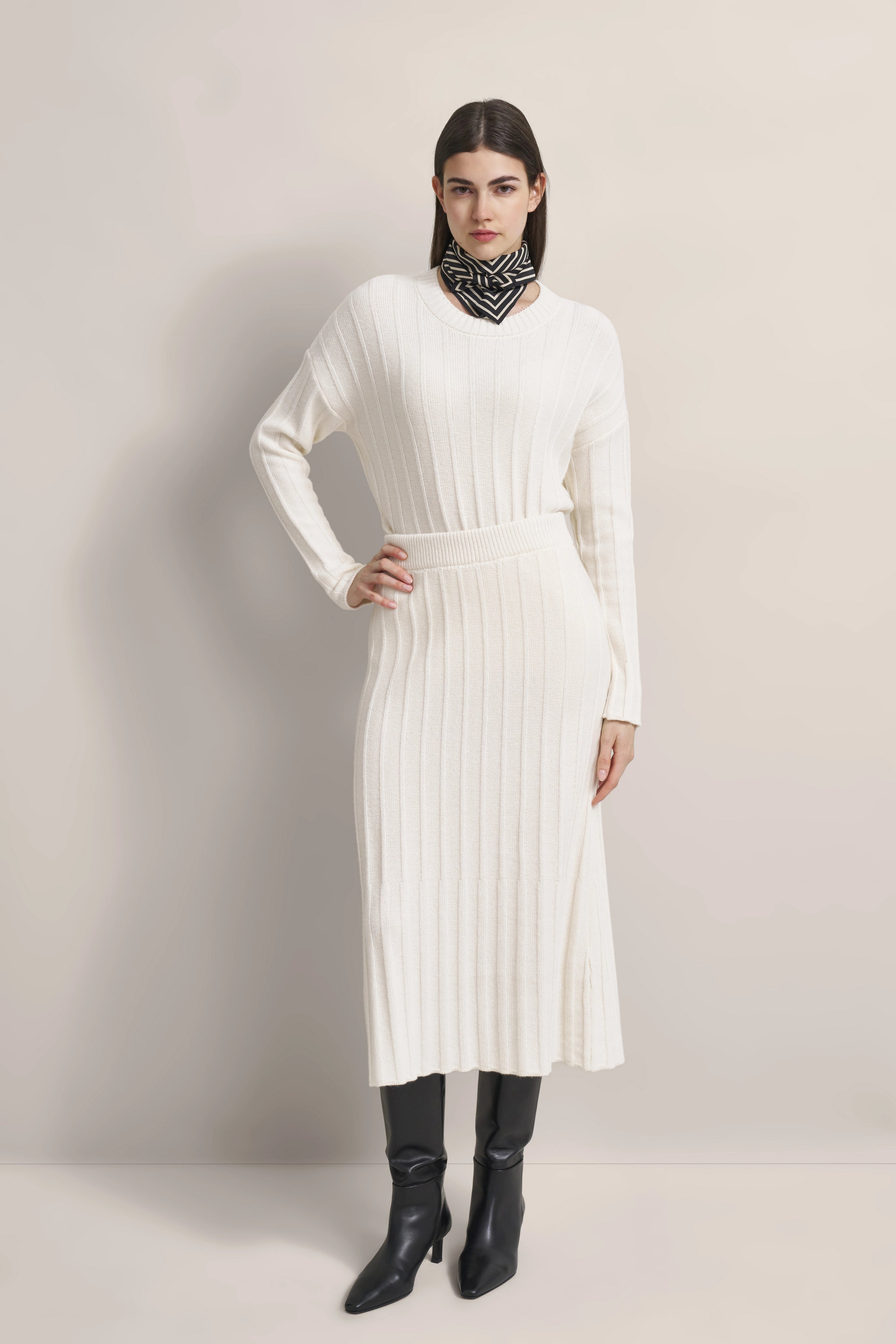 Modell in weißem, geripptem bugatti Strickkleid mit schwarzen Stiefeln und gemustertem Tuch, Hand in Taille.