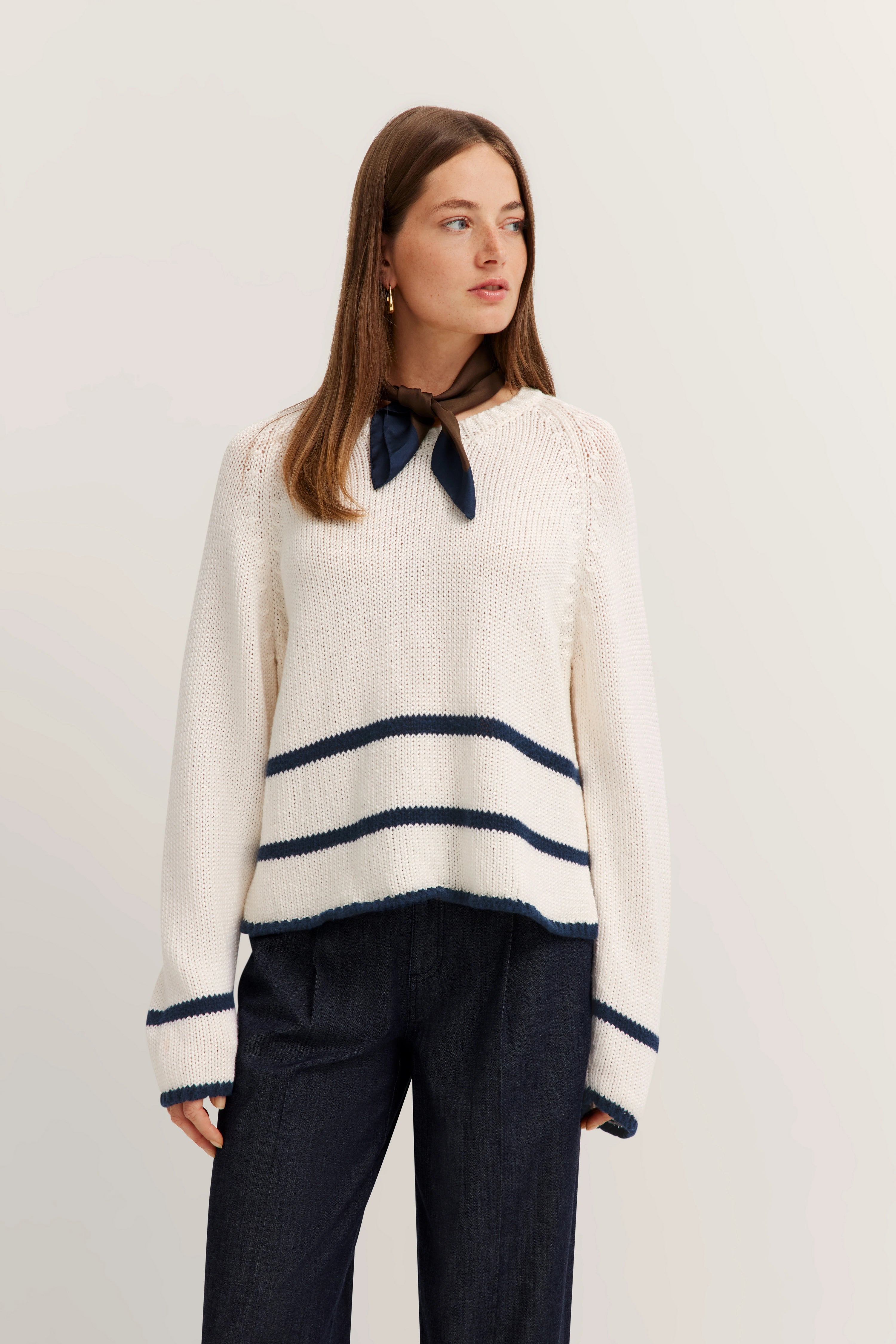 Weißer Strickpullover mit blauen Streifen, braunes Tuch, dunkle Hose. Modelbild, Blick zur Seite.