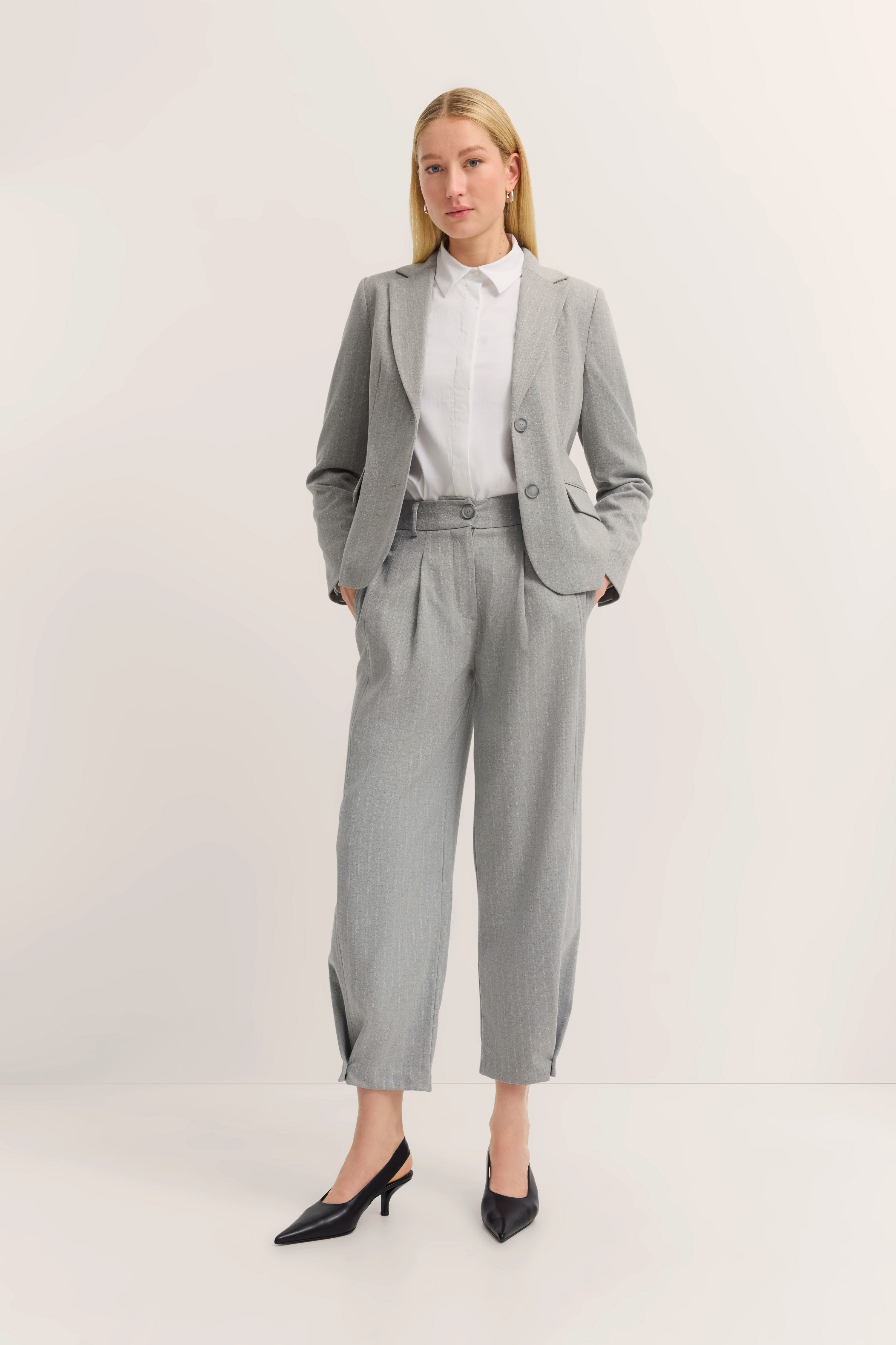 Grauer Anzug mit weißer Bluse, Modellbild, Hände in Taschen, elegantes Standing.