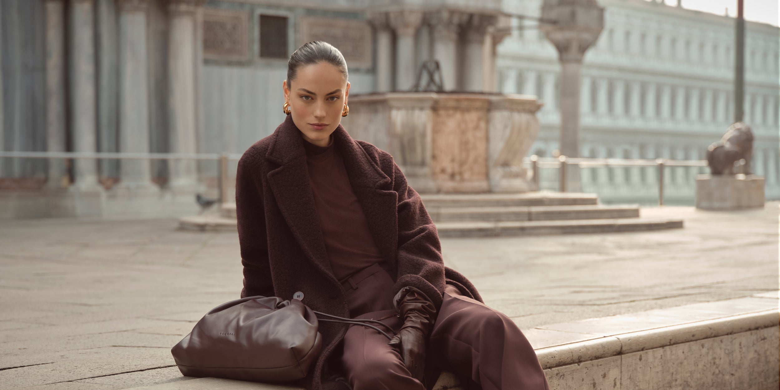 Eine Frau sitzt elegant auf einer steinernen Stufe, gekleidet in ein monochromes Outfit aus dunklem Bordeaux. Stilvoller, femininer Look mit urbaner Eleganz.