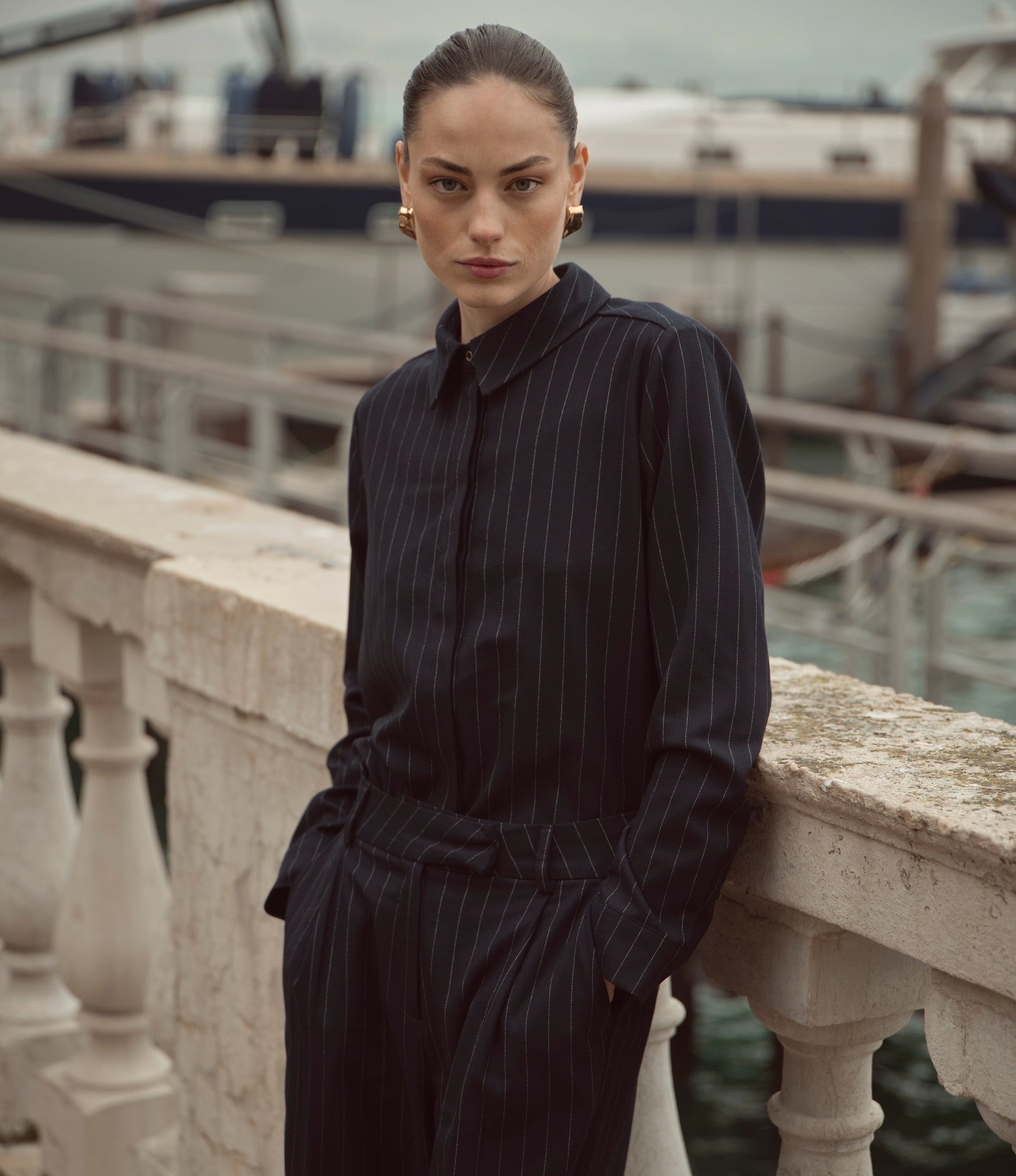 Eine Frau steht an einer steinernen Balustrade am Wasser und trägt einen dunkelblauen Nadelstreifenanzug. Der Look wirkt elegant, modern und selbstbewusst.