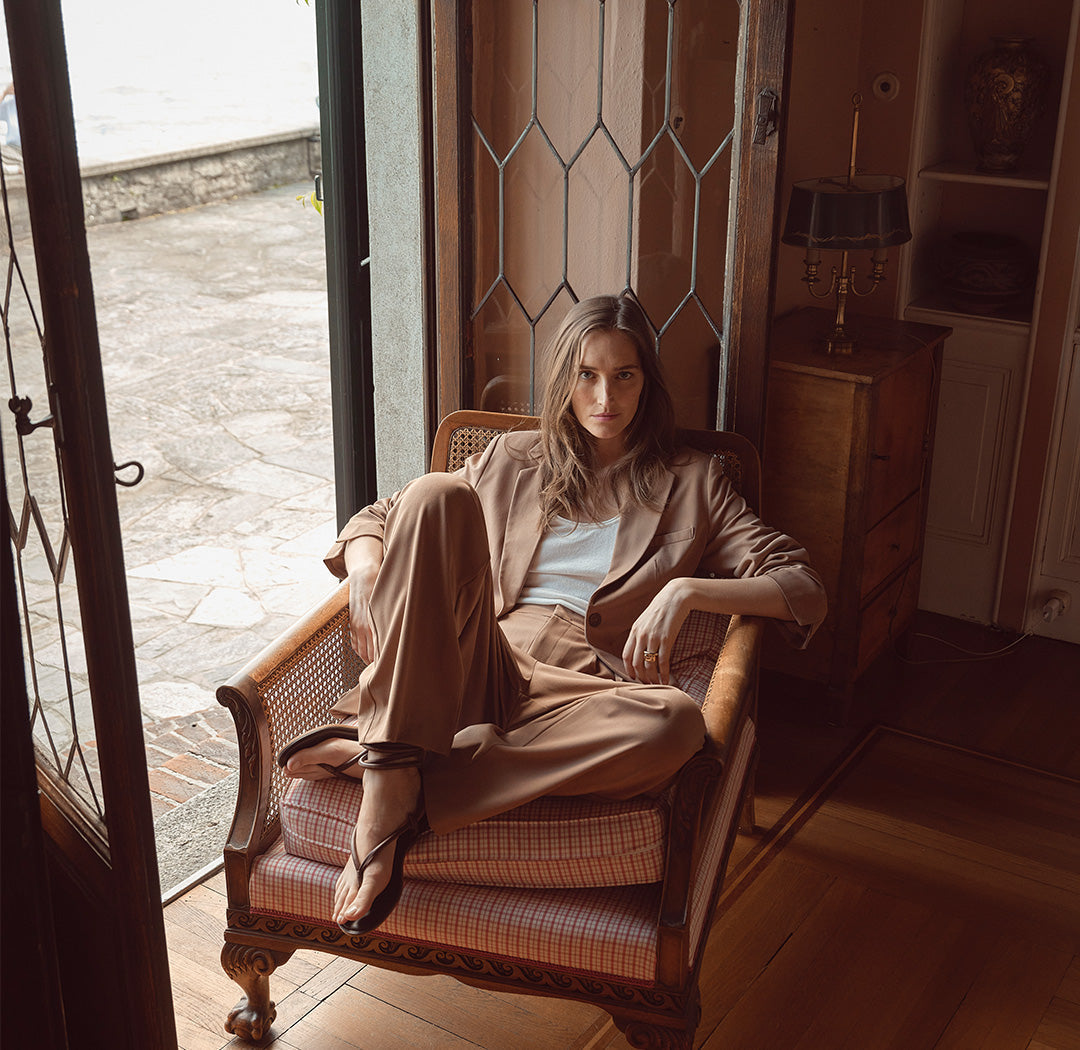 Eine Frau sitzt lässig in einem Sessel, trägt einen beigen Hosenanzug und ein helles Top. Der Look vereint entspannte Eleganz und modernen Chic.