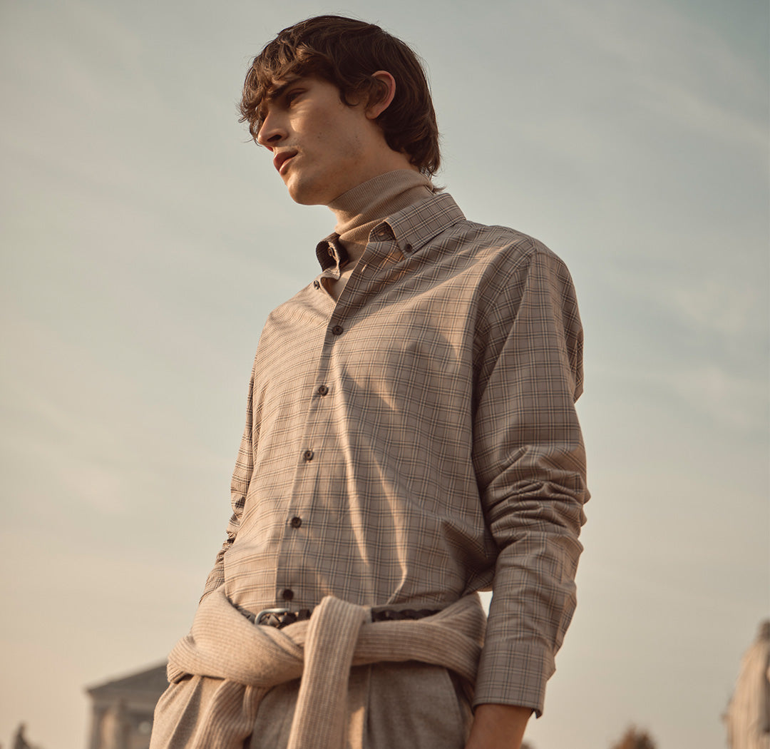 Ein Mann trägt ein kariertes Hemd über einem beigen Rollkragenpullover. Der Look wirkt elegant, herbstlich und zugleich lässig-modern im warmen Abendlicht.