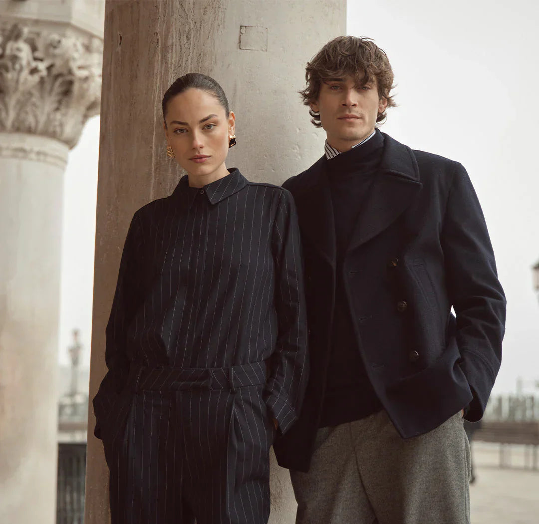 Ein stilvolles Paar steht vor einer steinernen Säule in einer eleganten, urbanen Umgebung.