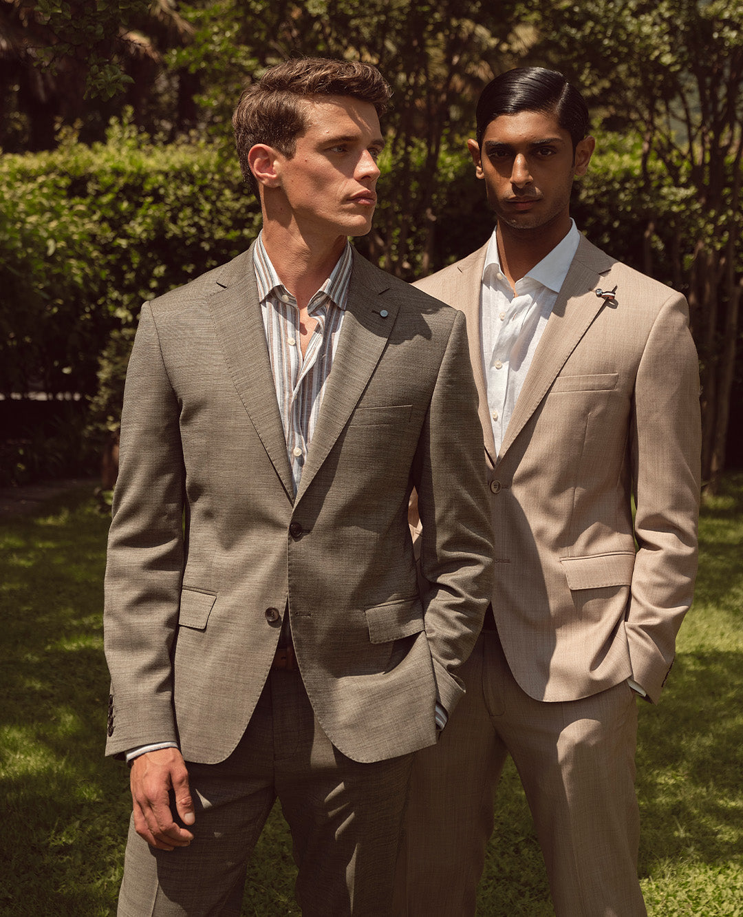 Zwei Männer tragen elegante Anzüge in hellen Naturtönen – der eine mit dezentem Streifenhemd, der andere mit klassischem weißen Hemd. Die Kombination aus sommerlichem Licht und edlen Stoffen unterstreicht die zeitlose Eleganz und Raffinesse der Looks.