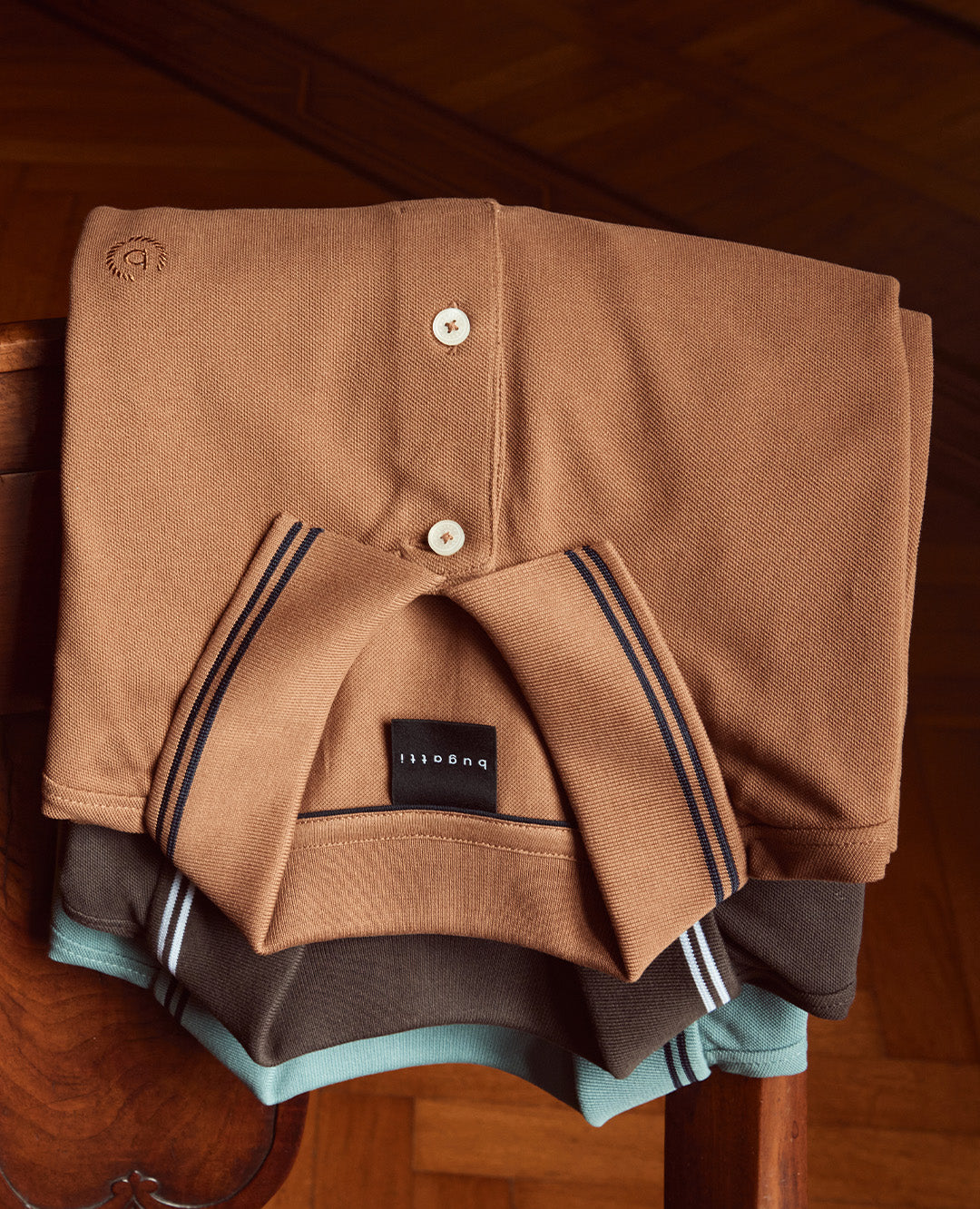 Drei sorgfältig gefaltete Poloshirts in Braun- und Blautönen liegen auf einem Holzstuhl. Hochwertige Materialien und dezente Details betonen eleganten Stil.