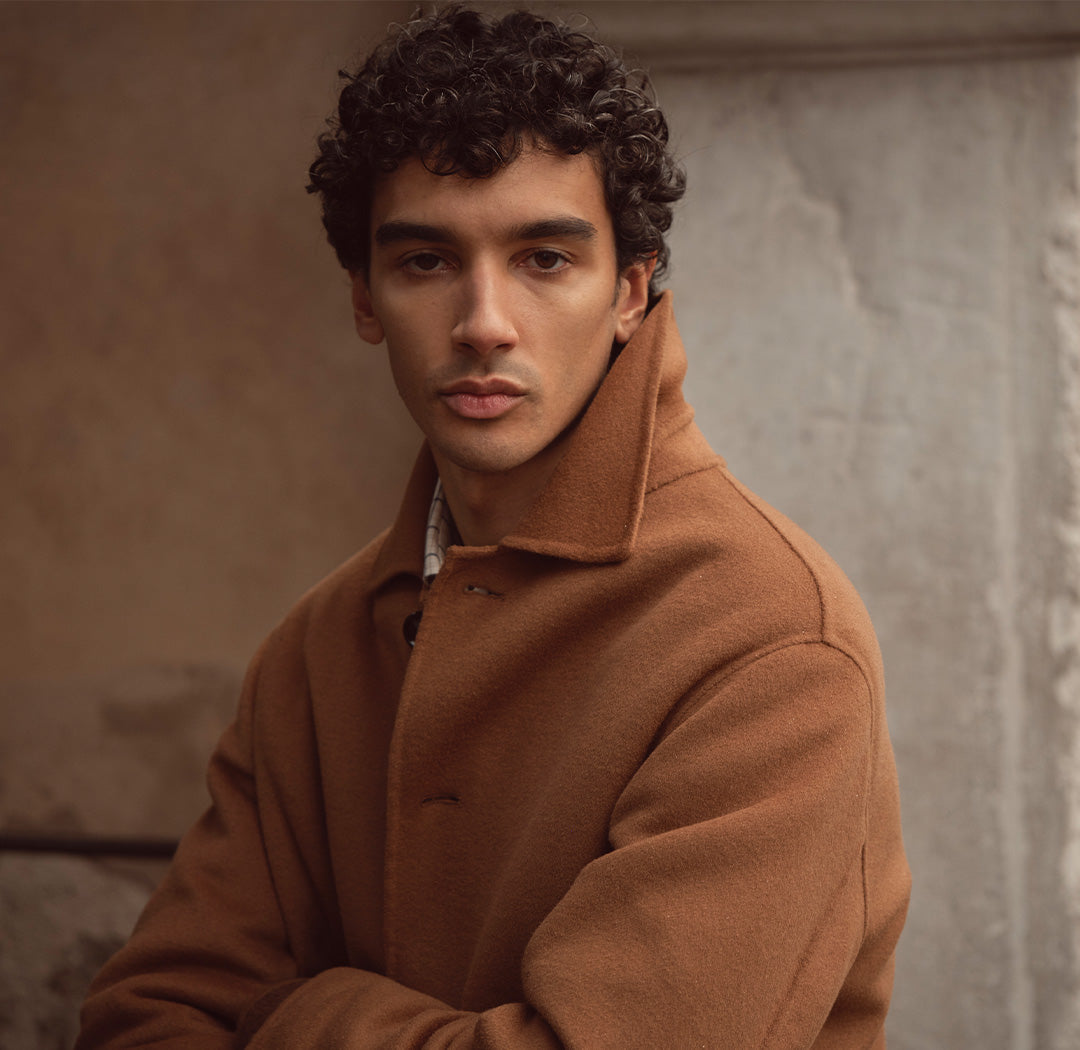 Ein Mann trägt einen eleganten, camelbraunen Mantel und blickt ruhig in die Kamera. Der Look wirkt klassisch, selbstbewusst und stilvoll zeitlos.