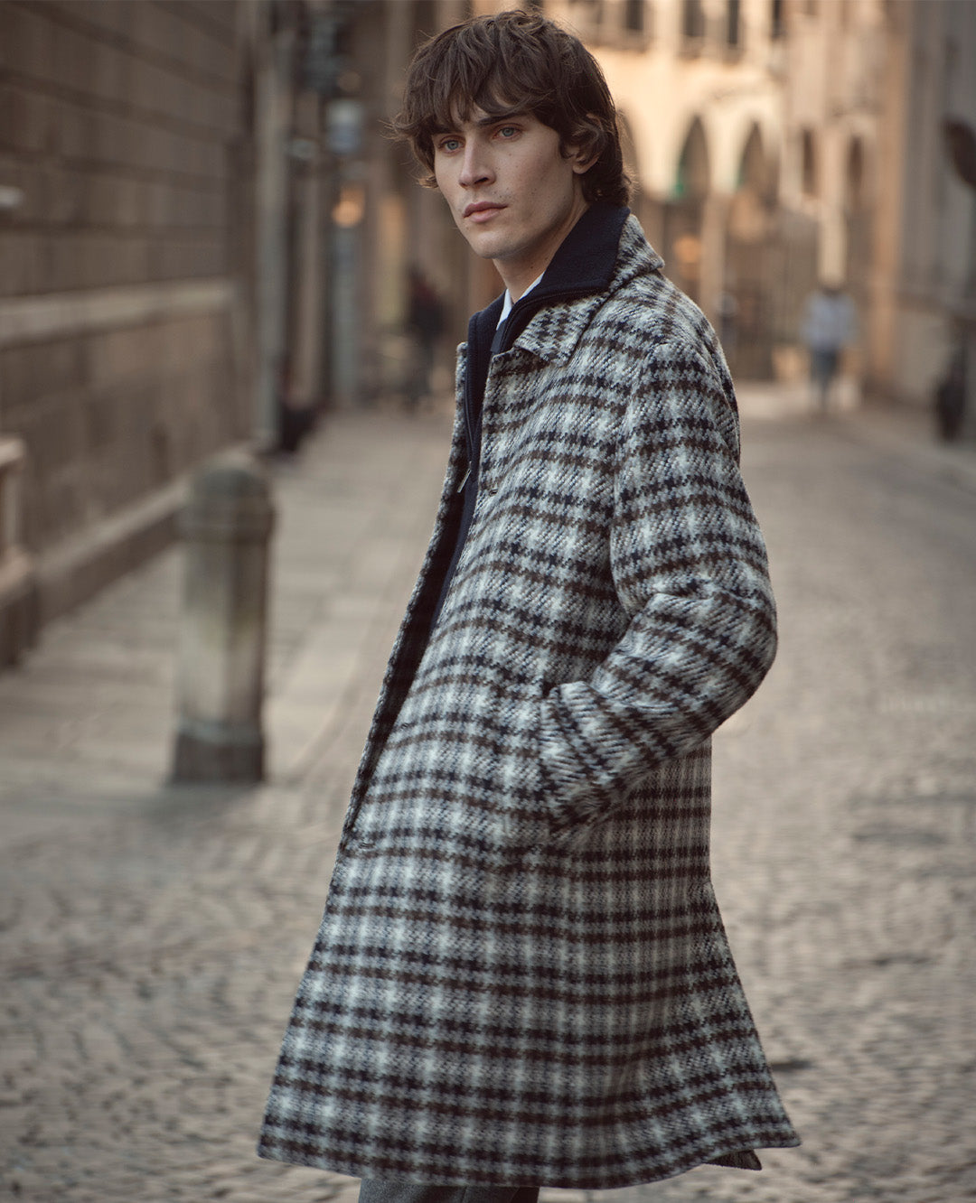 Ein Mann steht auf einer gepflasterten Straße und trägt einen langen karierten Mantel in gedeckten Tönen. Sein Blick wirkt ruhig und stilvoll urban.