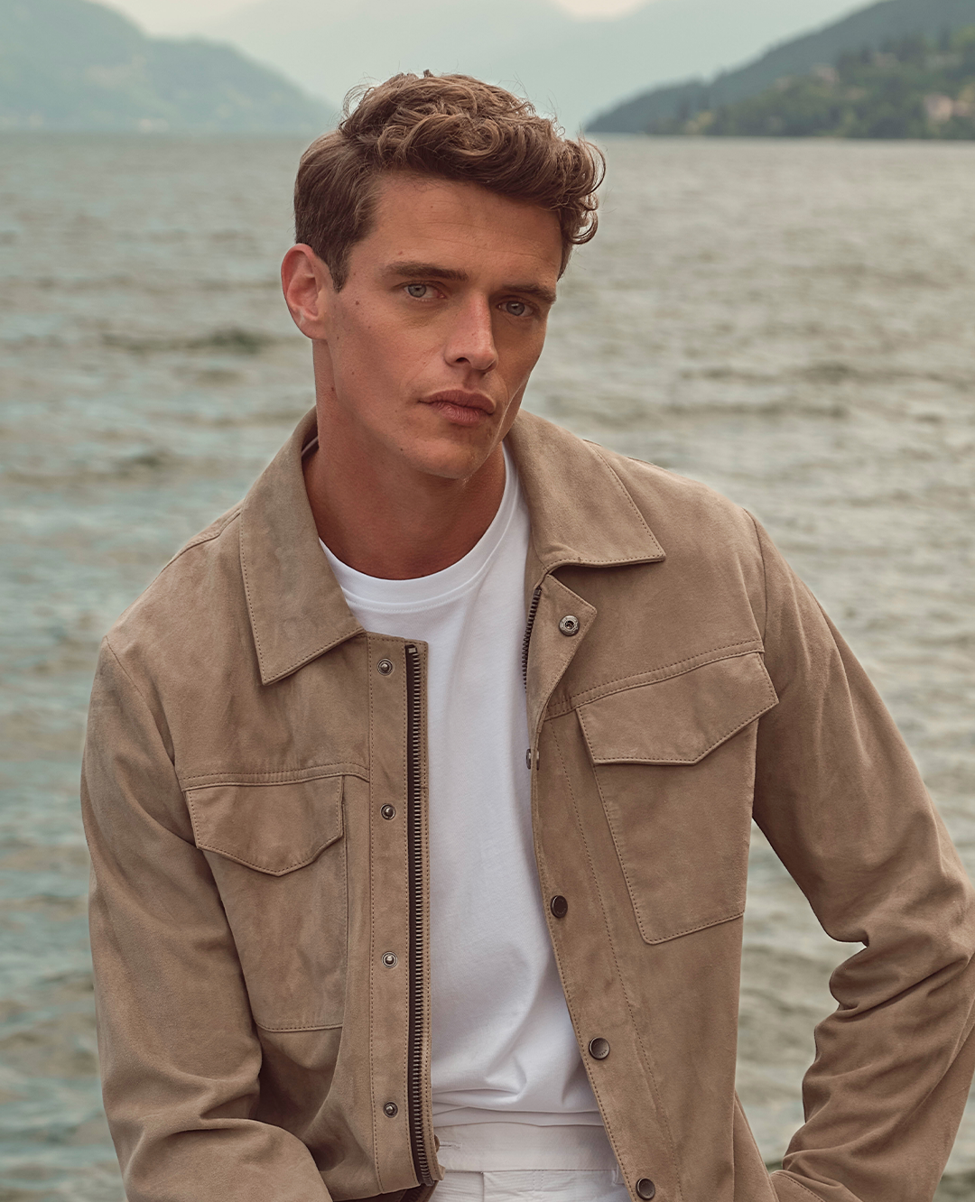 Ein Mann trägt eine beigefarbene Wildlederjacke über einem weißen T-Shirt. Der Look wirkt lässig, modern und elegant vor der Kulisse eines Sees.