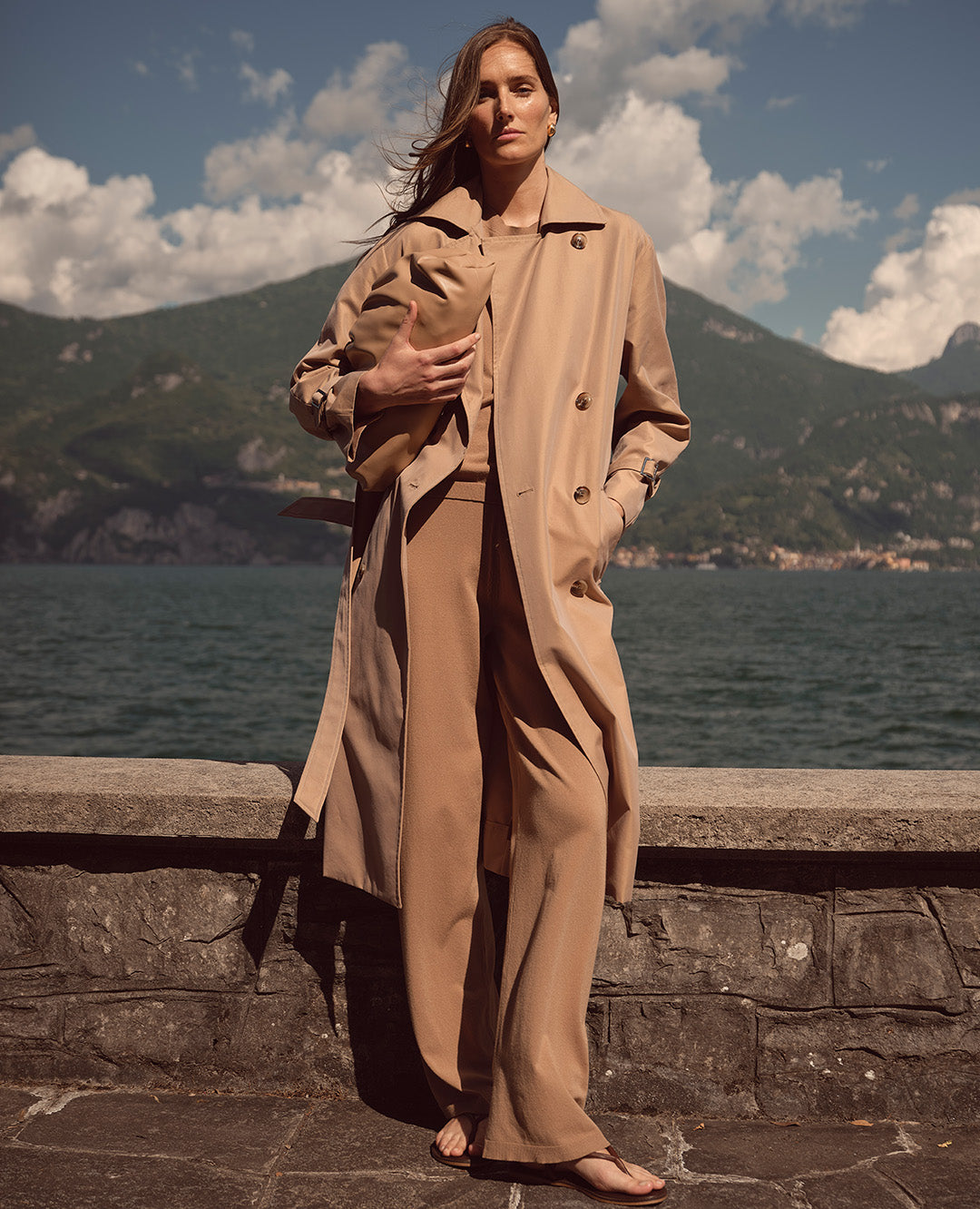 Eine Frau steht vor einer malerischen Seelandschaft und trägt ein hellbraunes Ensemble mit Trenchcoat. Der Look wirkt elegant, natürlich und selbstbewusst.