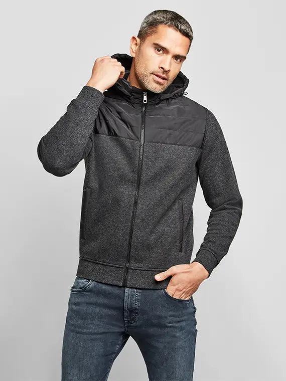 Ein Mann trägt eine dunkelgraue Kapuzenjacke mit schwarzer Stepppasse und Jeans. Der Look wirkt sportlich, modern und gleichzeitig lässig elegant.