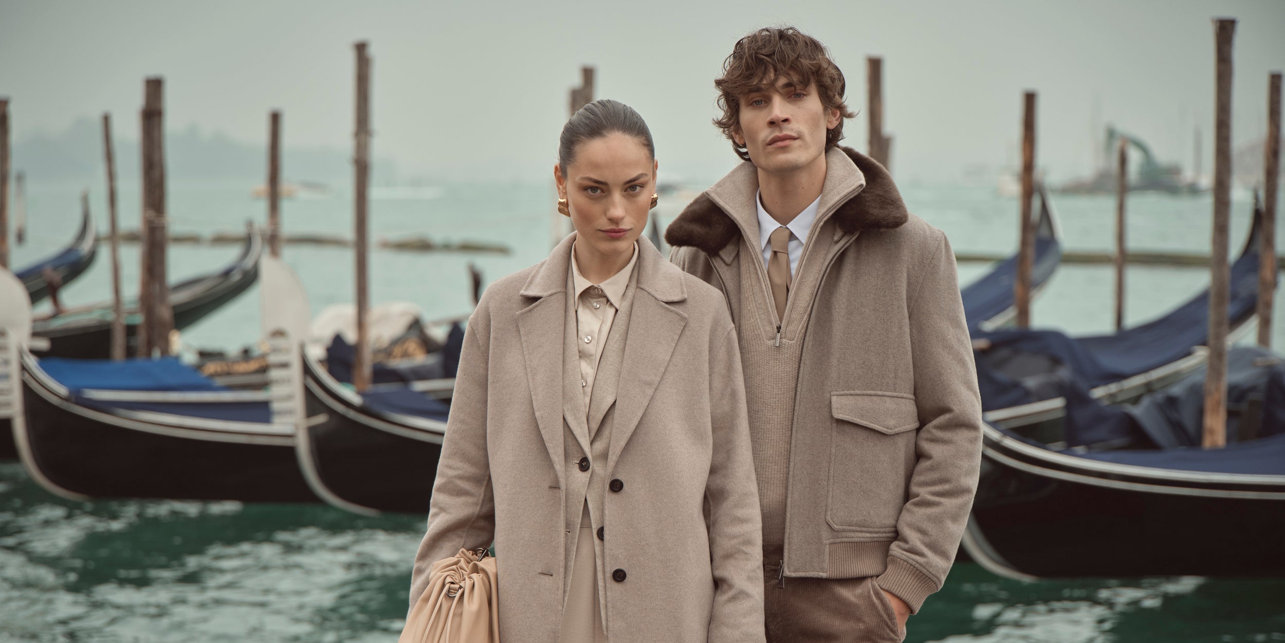 Frau und Mann tragen beigefarbene bugatti Mäntel aus der Herbst/Winter-Kollektion 2025, fotografiert vor Gondeln in Venedig – elegante Womenswear und Menswear im Venetian Chic.