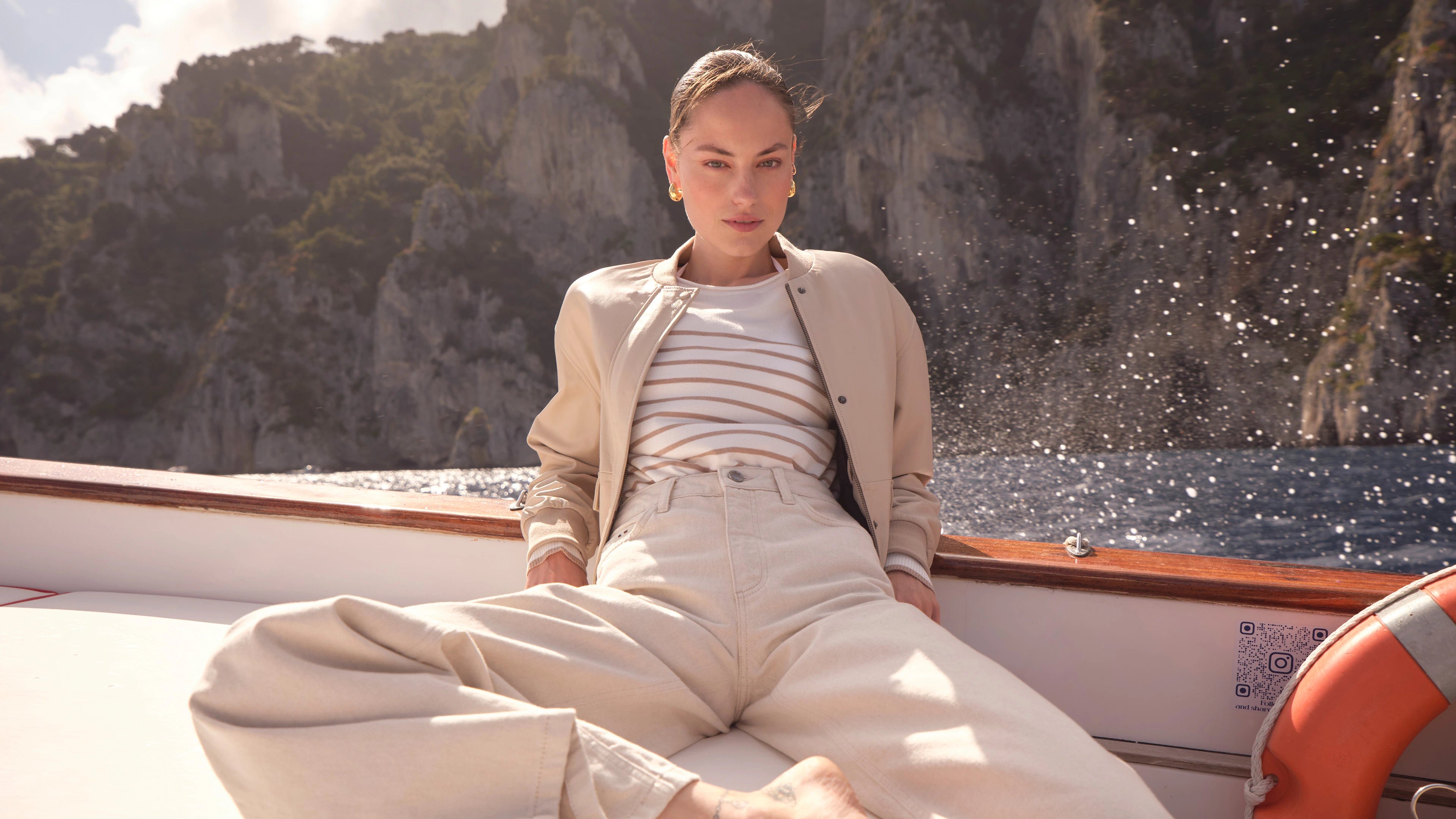 Person wearing light beige jacket and striped shirt sitting on boat with rocky cliffs and water in background
