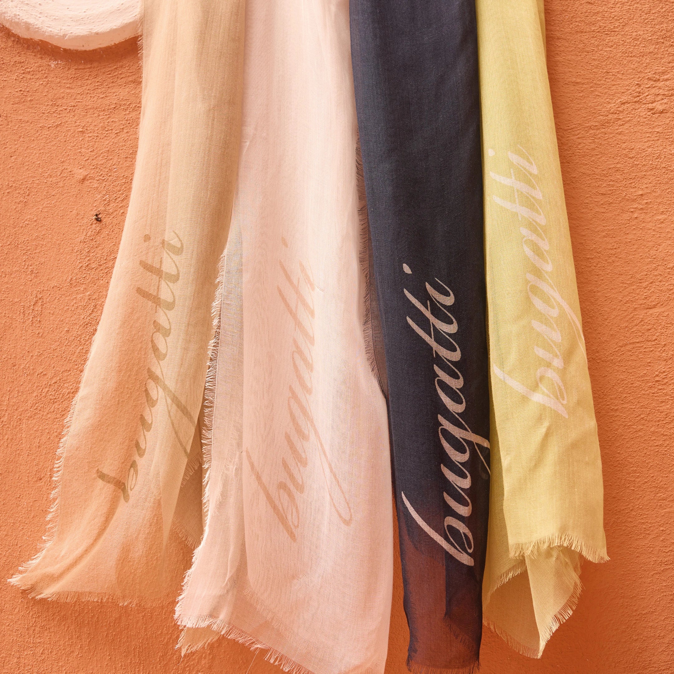 Four lightweight scarves in soft peach, cream, dark blue, and pale yellow hanging against a textured terracotta wall with "bugatti" branding