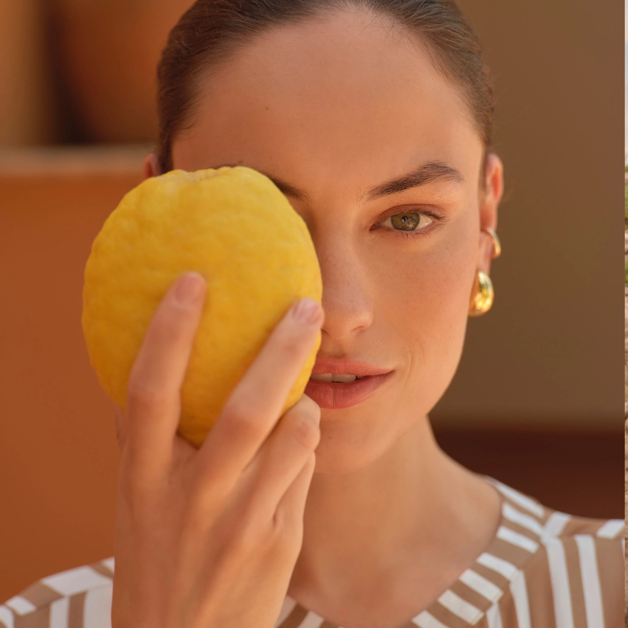 Woman with pulled-back hair holding large yellow citrus fruit over one eye wearing gold hoop earrings and striped top
