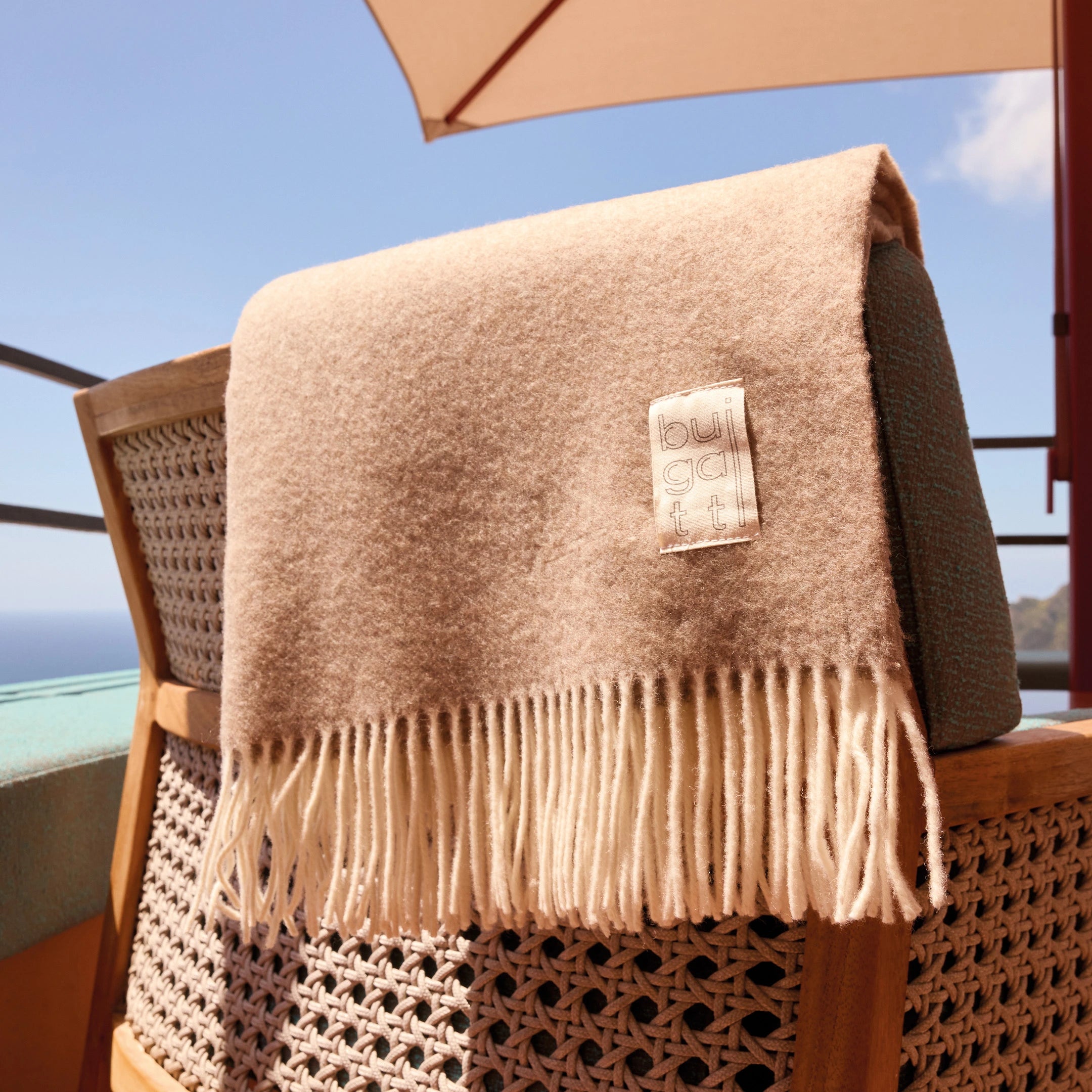 Beige fringed woolen scarf draped over woven chair on balcony with blue sky and ocean in background