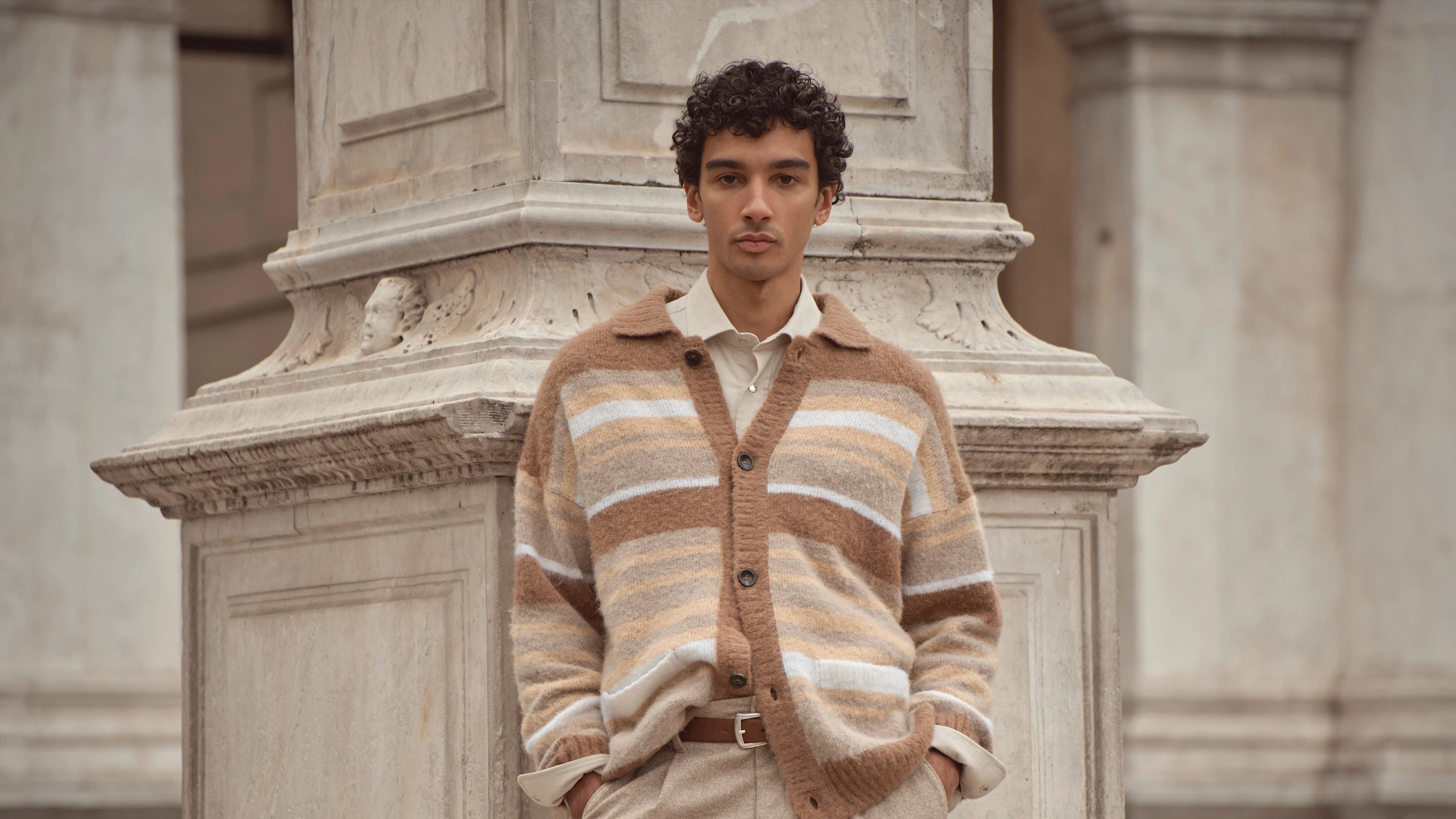 Mann trägt beigefarbenen Strick-Cardigan mit Streifenmuster aus der bugatti Herbst/Winter-Kollektion 2025 – lässiger Style in Naturtönen, kombiniert mit Hemd und heller Hose vor venezianischer Architektur.