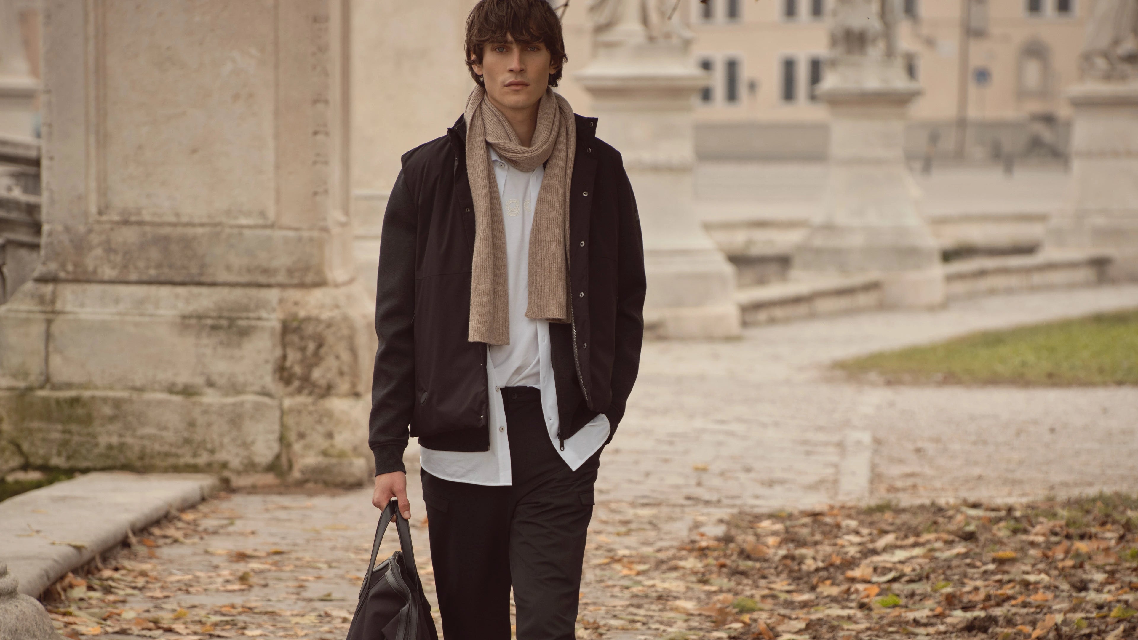 Mann in schwarzer Jacke, weißem Hemd und beigefarbenem Schal aus der bugatti Herbst/Winter-Kollektion 2025 geht durch eine historische Kulisse in Venedig – moderner City-Look mit sportlich-elegantem Akzent.