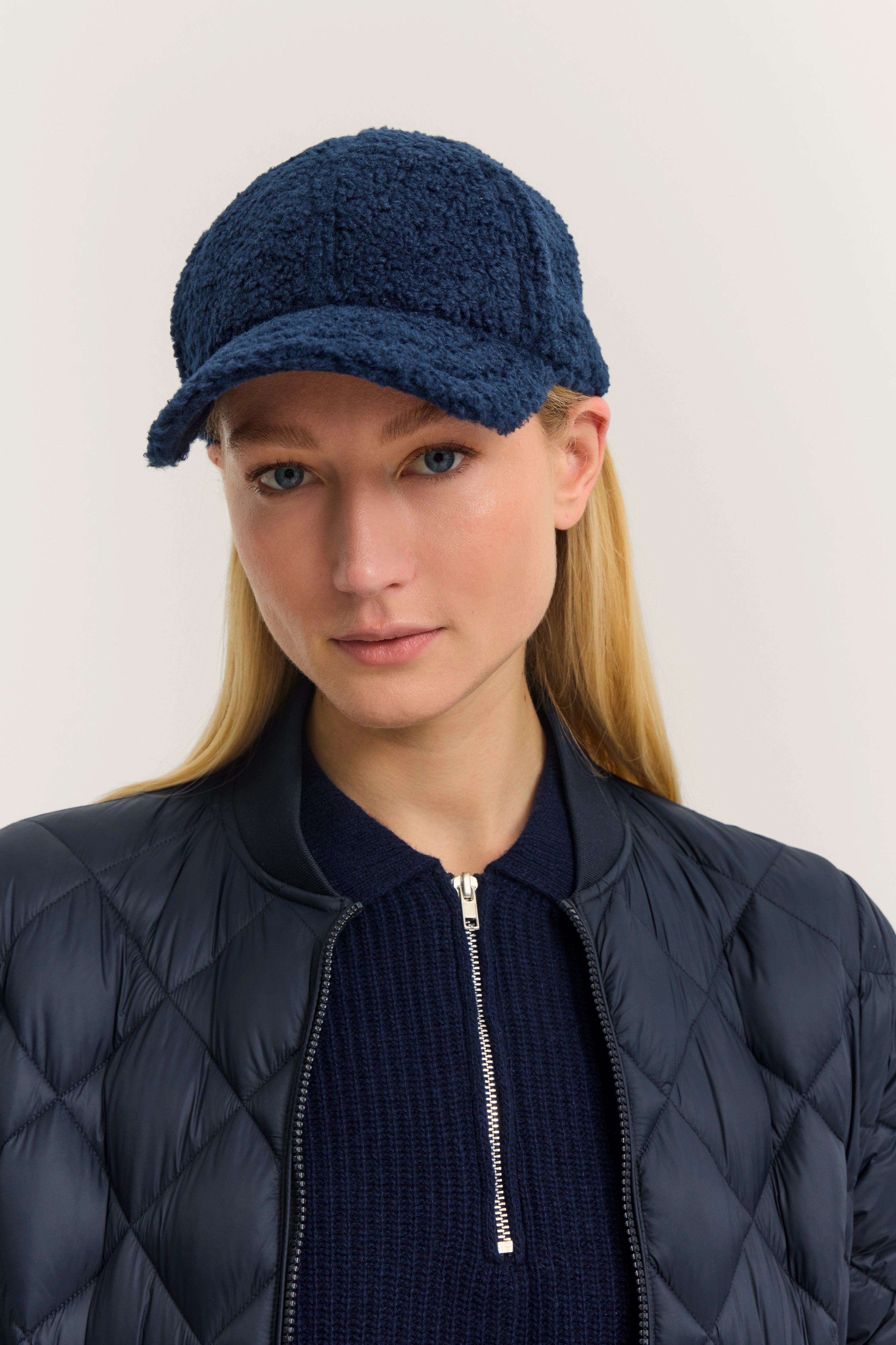 Model trägt blaue Steppjacke, dunkelblauen Pullover und Mütze. Nahaufnahme, frontal.