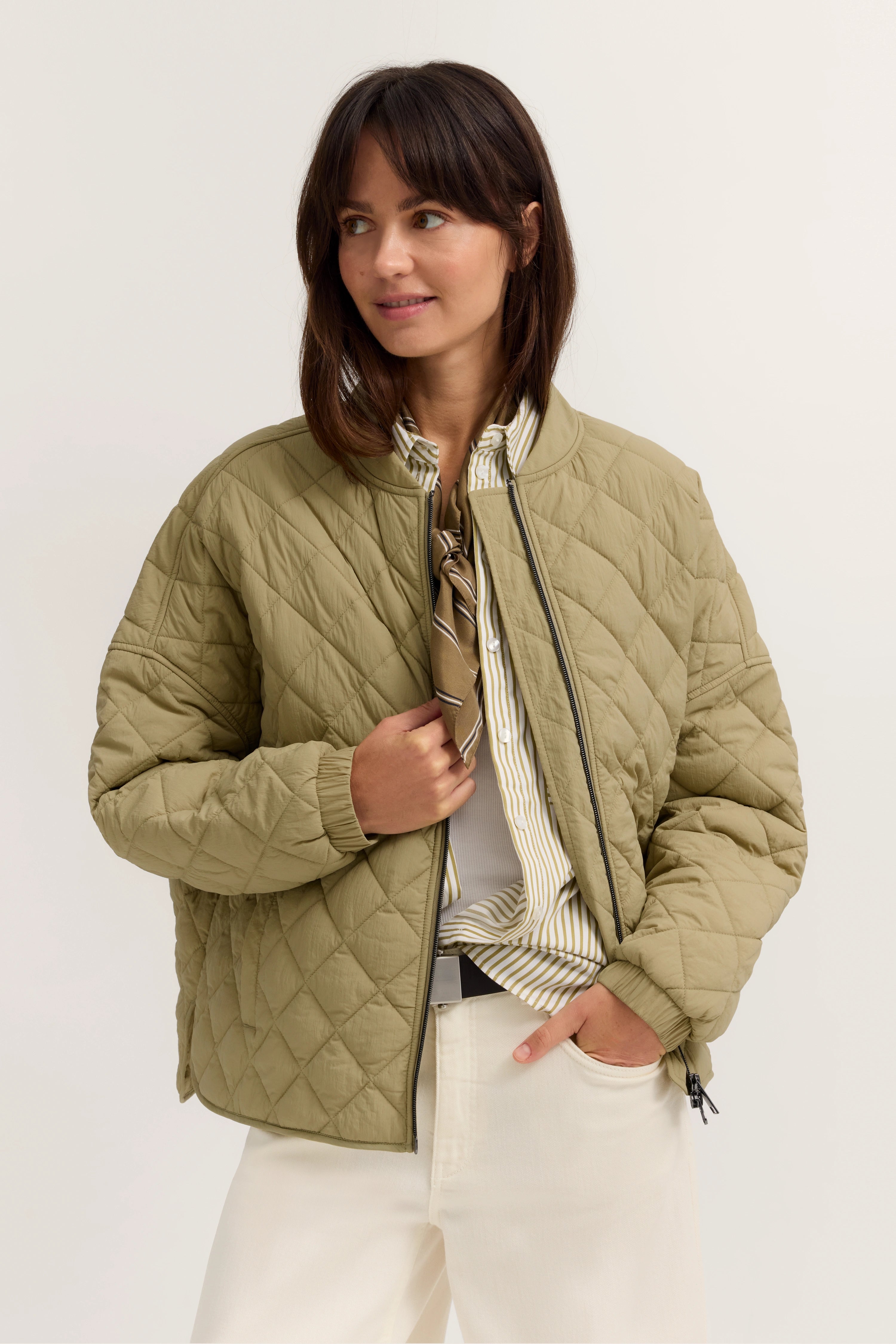 Model trägt gesteppte, olivgrüne bugatti Jacke mit gestreiftem Hemd und weißer Hose, legeres Posing.